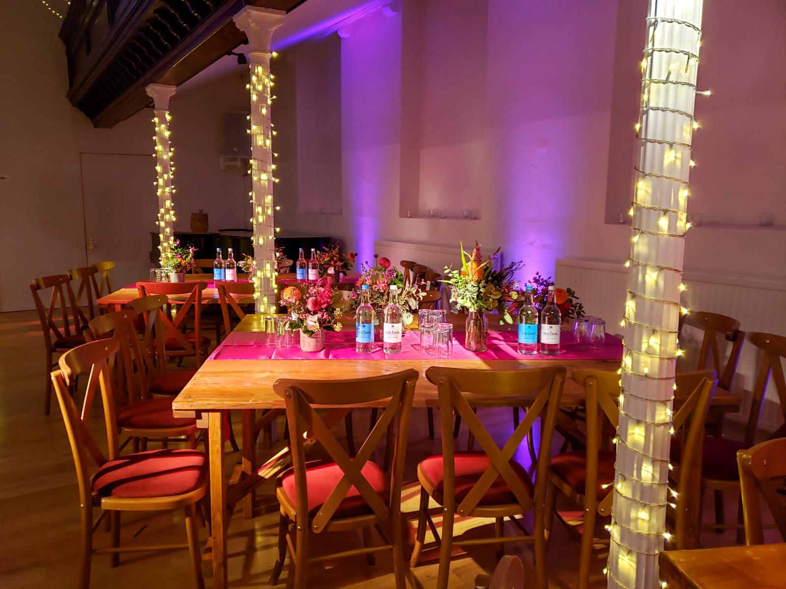 An event venue with wooden tables, red cushioned chairs, floral centerpieces, and columns wrapped in string lights at The Amadeus.