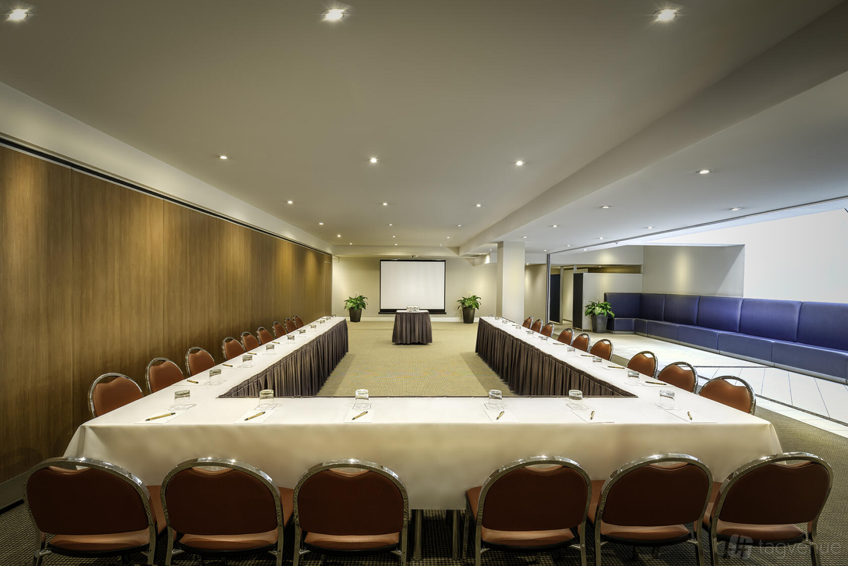A hotel conference room with U-shaped tables, red chairs, a projection screen, and wood-paneled walls at Batman's Hill on Collins.