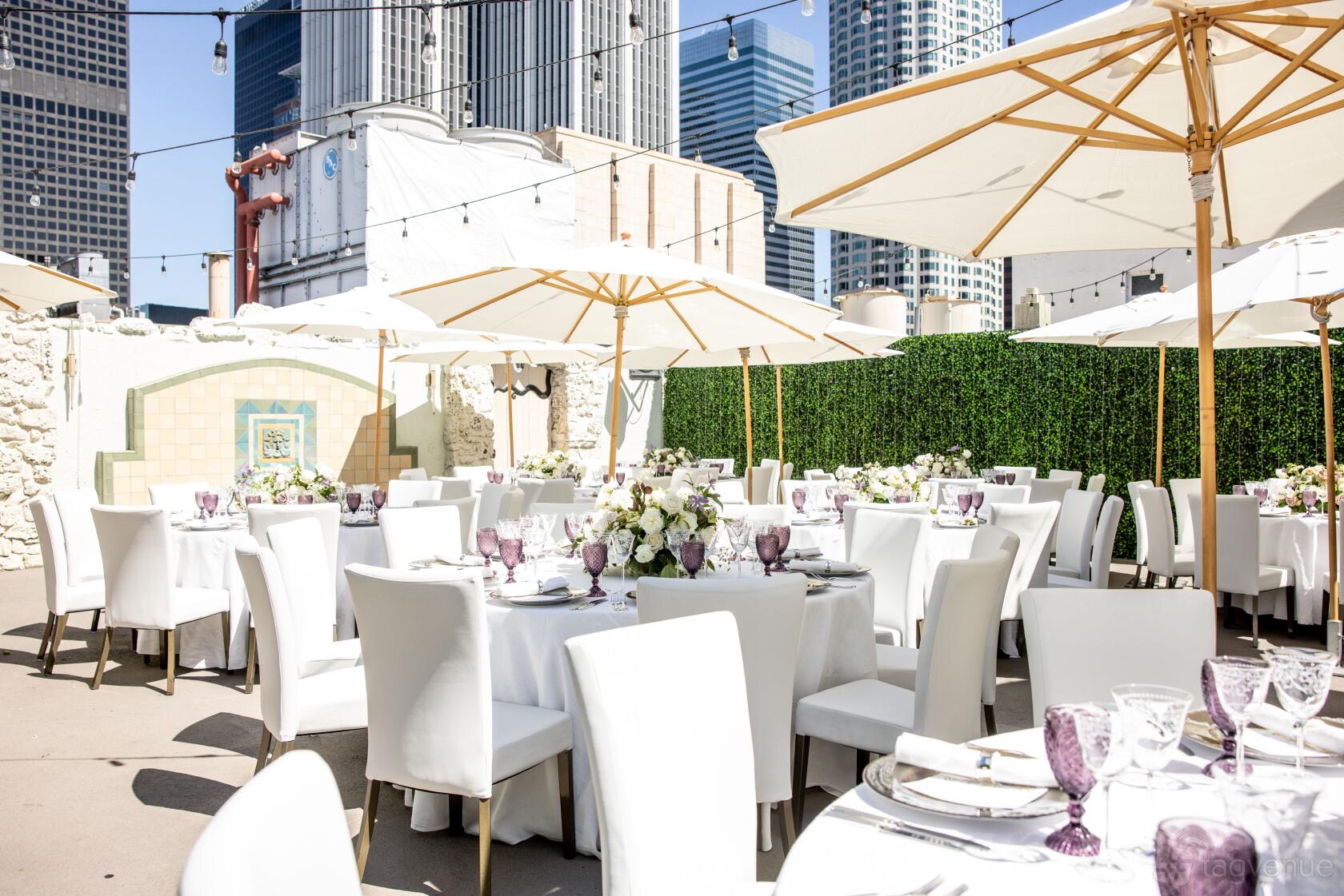 An outdoor event space with round tables draped in white linens, umbrellas, and city views at The Oviatt: Penthouse & Event Deck.
