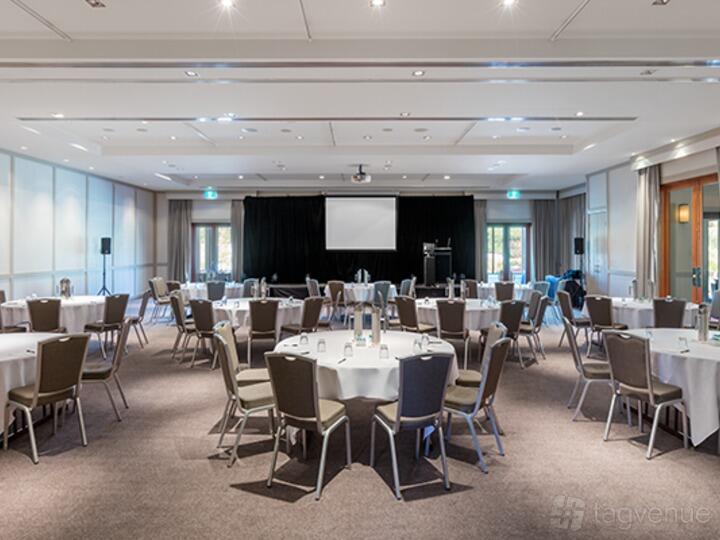 A function room with round tables, brown chairs, and a stage with a projector screen at Yarra Valley Lodge.