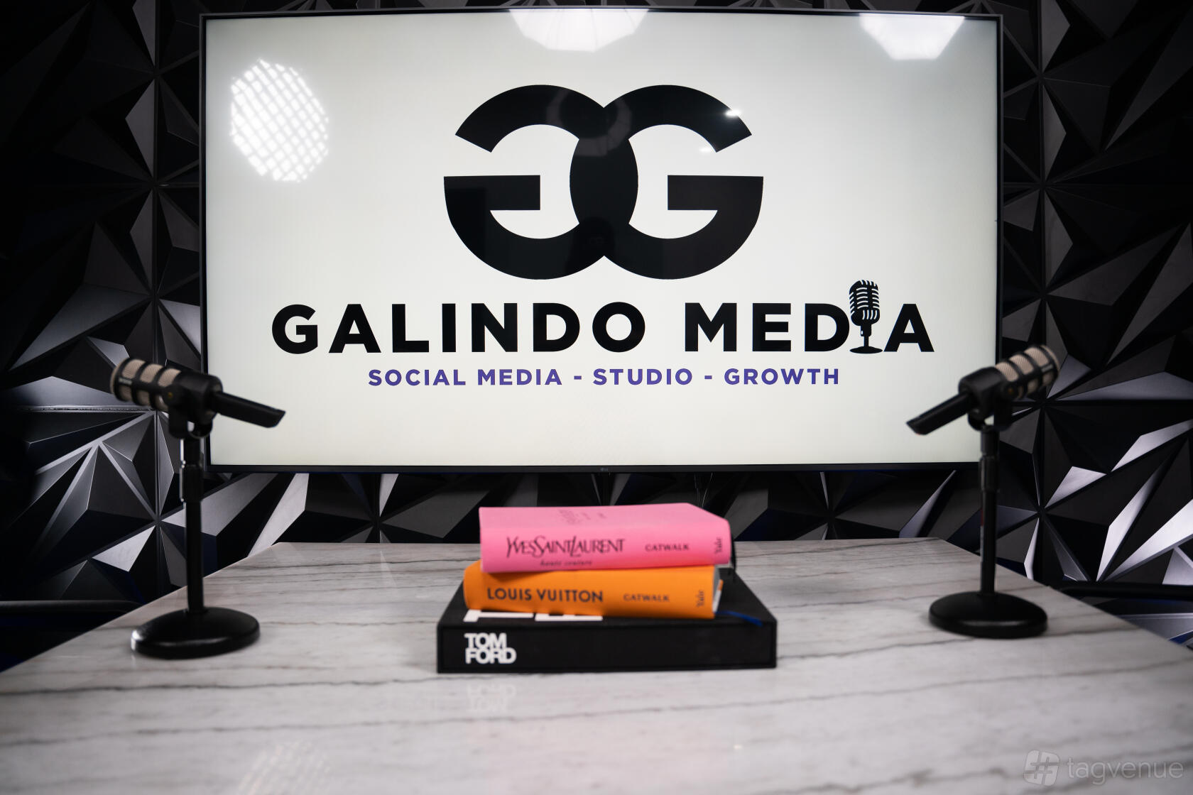 A podcast studio with tabletop microphones, designer books, and a large screen displaying the Galindo Media Studio logo.