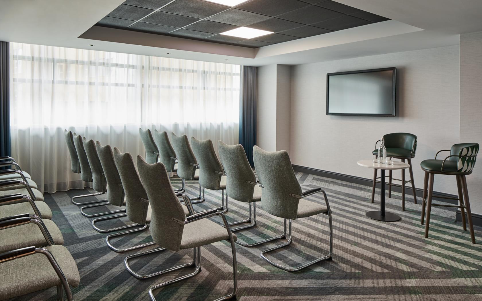 A hotel meeting room with rows of modern grey chairs, a wall-mounted screen, and floor-to-ceiling windows at Manchester Marriott Hotel Piccadilly.