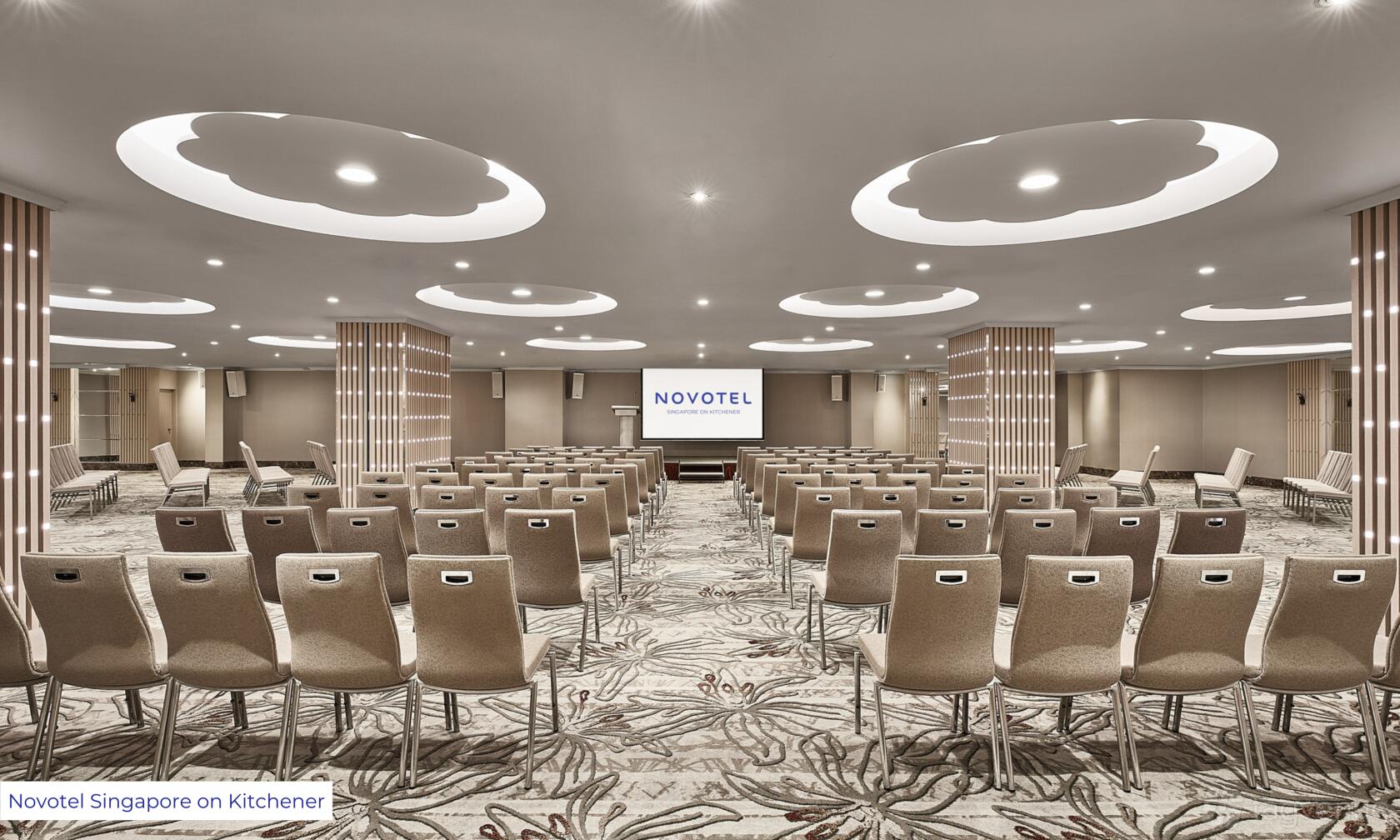 A banqueting hall with rows of beige chairs, patterned carpeting, and ceiling lights at CALI, Novotel on Kitchener Road.
