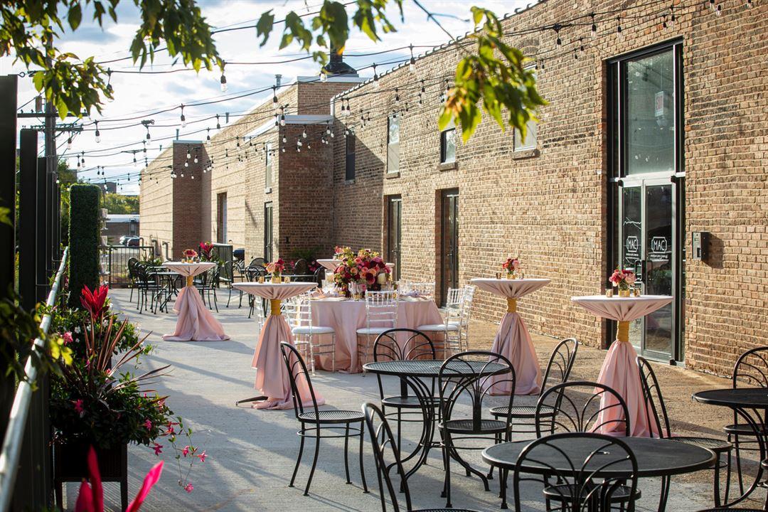 An outdoor event space with exposed brick walls, string lights, round metal tables, and pink-clothed cocktail tables at Morgan Arts Complex.