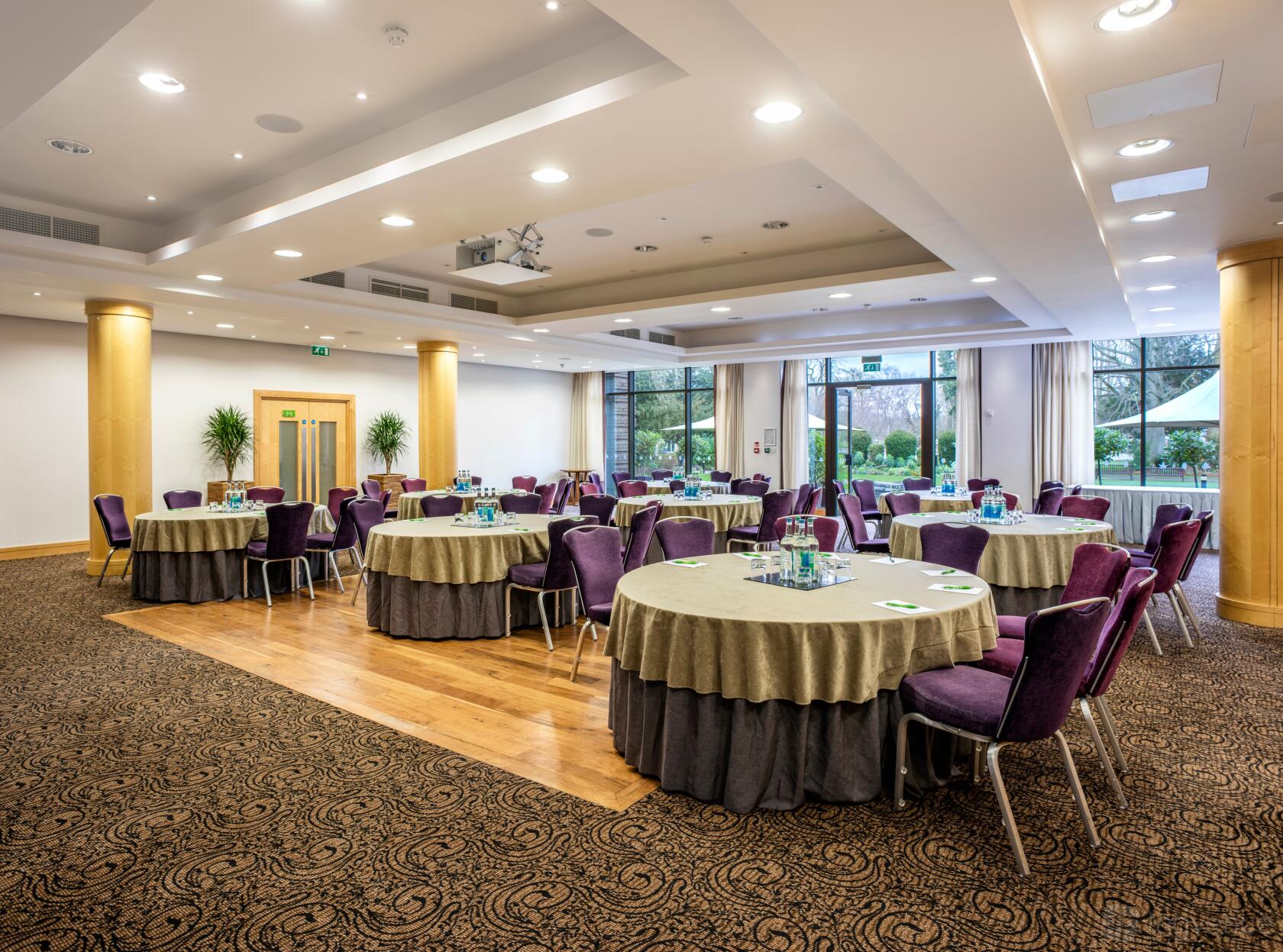 An event space with round tables draped in grey linens, purple chairs, and large windows overlooking greenery at The Lensbury.