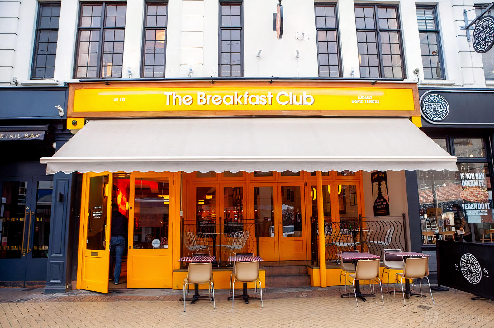 A café with a bright yellow facade, large windows, and outdoor seating at The Breakfast Club Chelmsford.