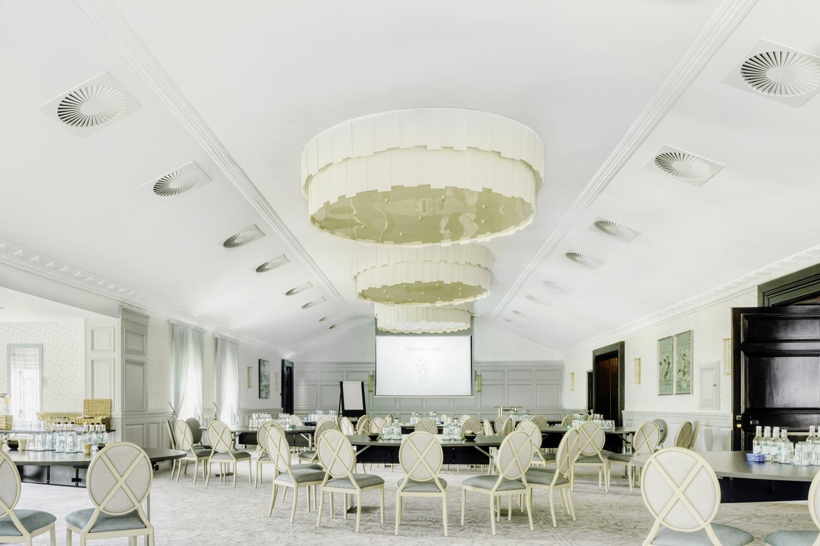 A spacious hotel conference room with round tables, upholstered chairs, large chandeliers, and a projection screen at Pennyhill Park.