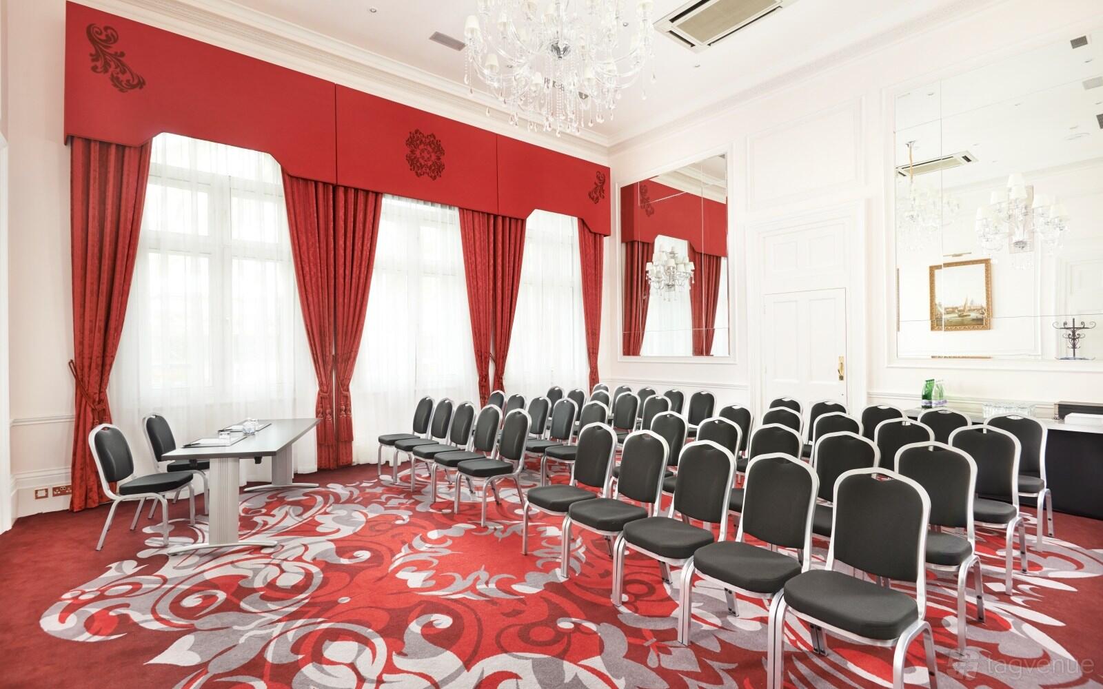 A meeting room with red patterned carpet, tall windows with red drapes, and rows of black chairs at Clermont Charing Cross.
