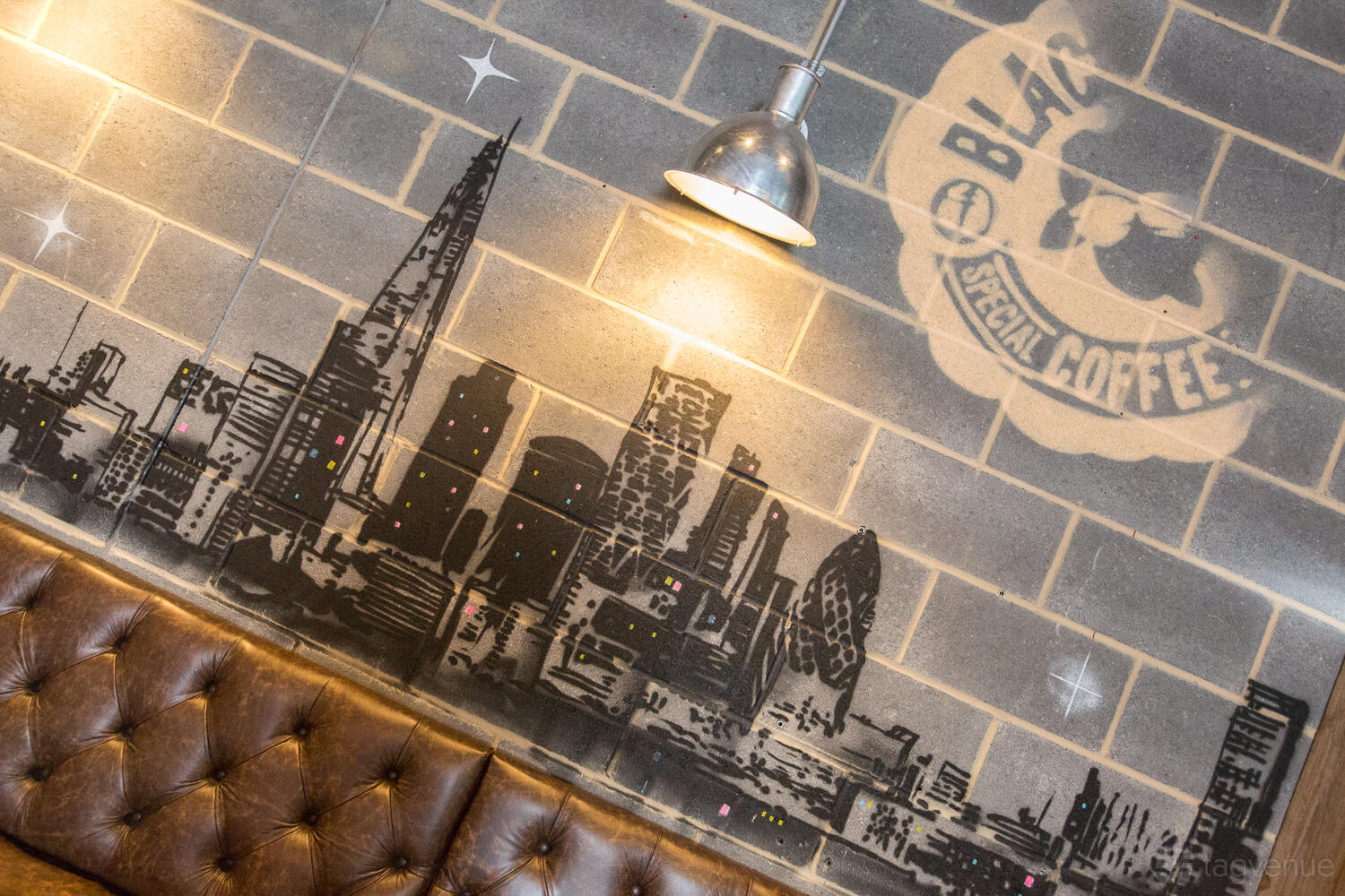 A café with a leather bench, exposed brick wall, city skyline mural, and industrial pendant light at Black Sheep Coffee Southbank Tower.