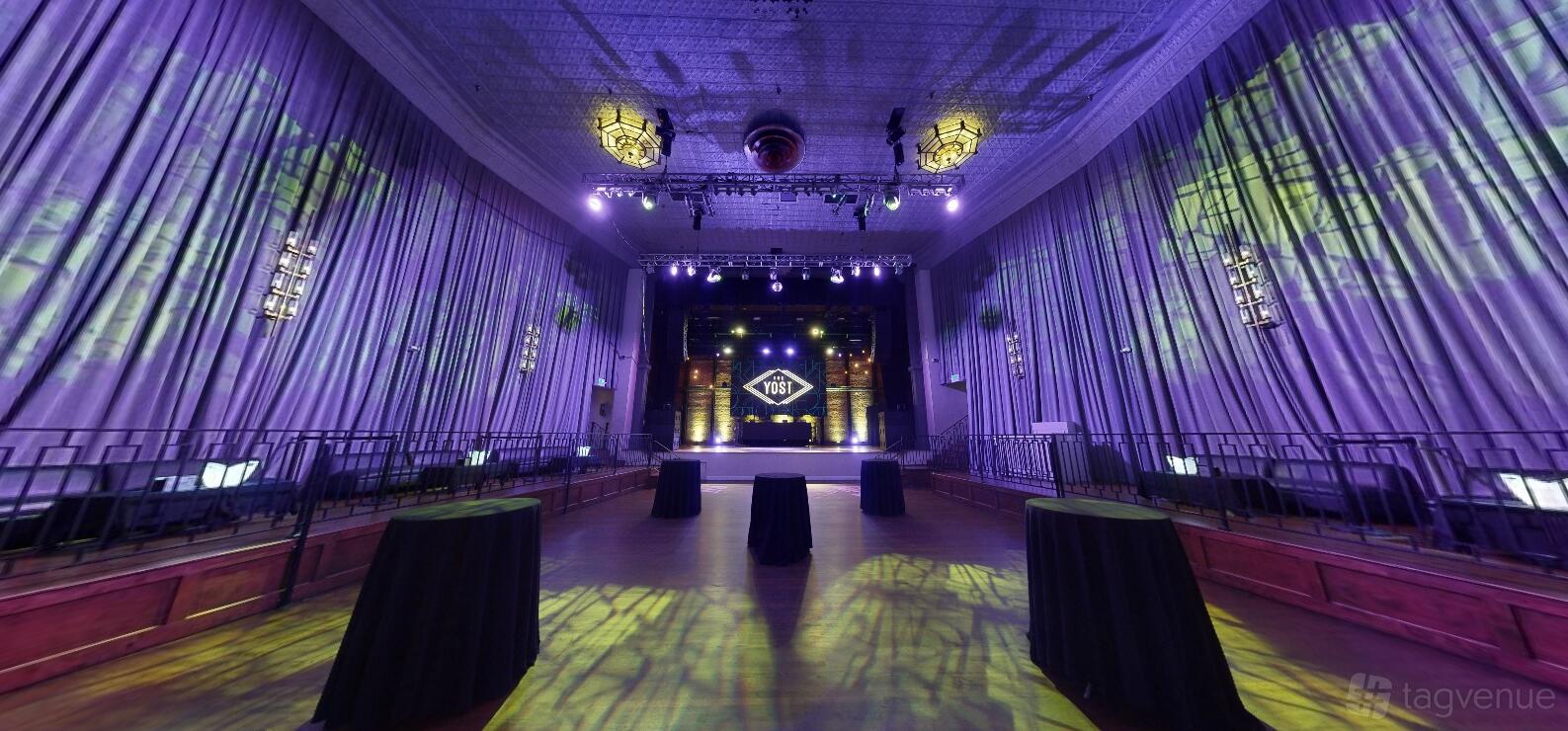 A theatre with a wide stage, dramatic uplighting, and high ceilings draped in floor-to-ceiling curtains at The Yost Theater.