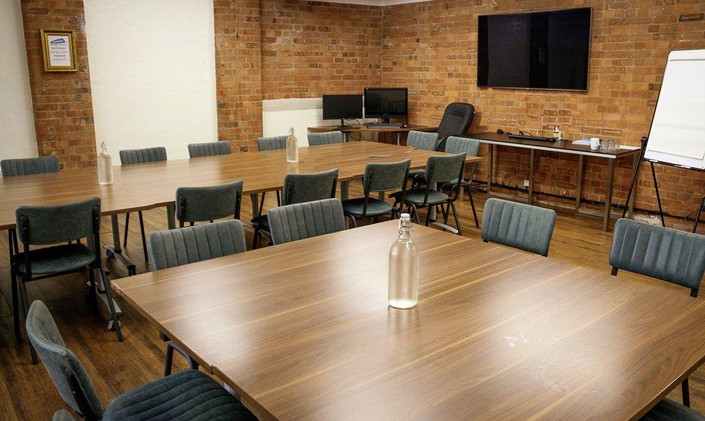 A meeting room with exposed brick walls, wood tables, green chairs, and a wall-mounted screen at Remark! Events.