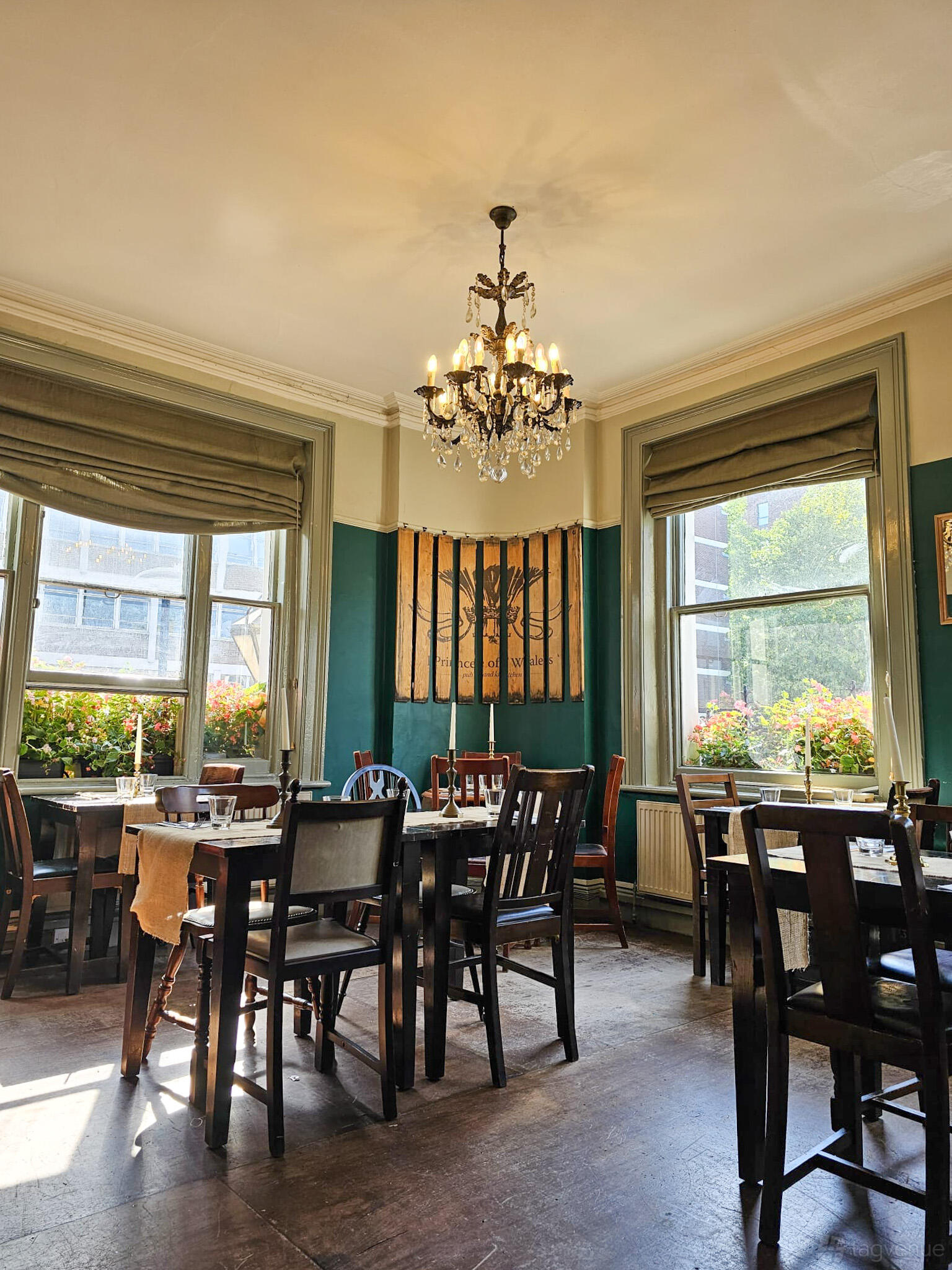 A pub dining room with large windows, wooden tables and chairs, a chandelier, and green accents at The Prince of Wales.