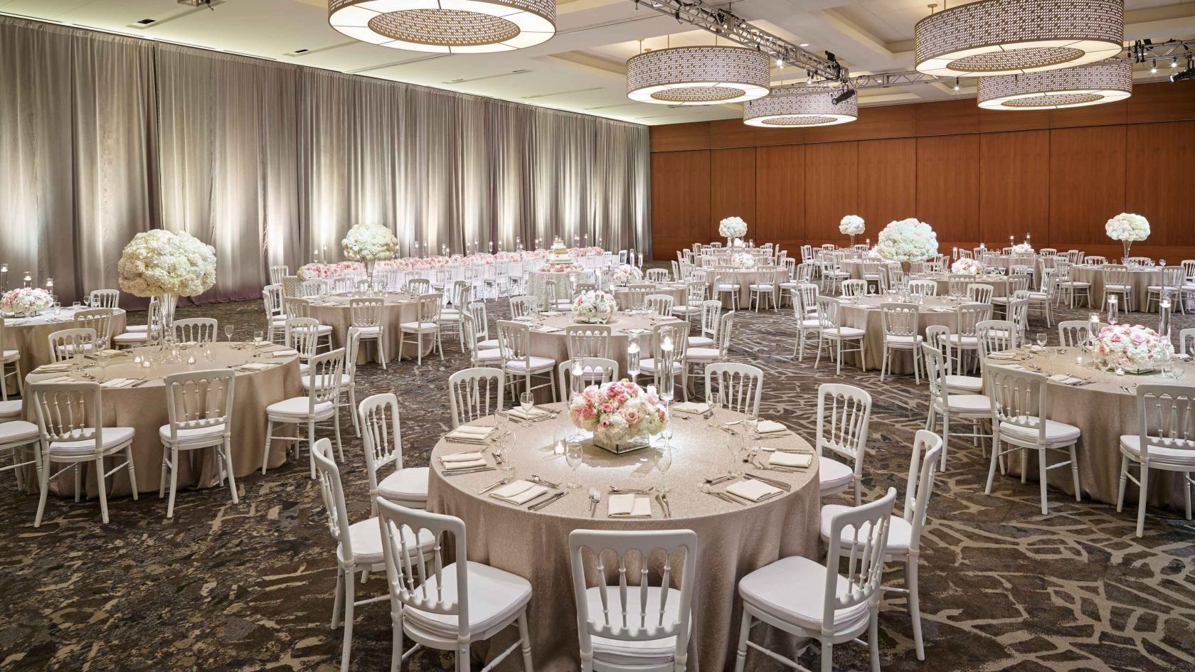 A hotel event space with round tables, white chairs, floral centerpieces, and overhead chandeliers at The Westin Galleria Dallas.