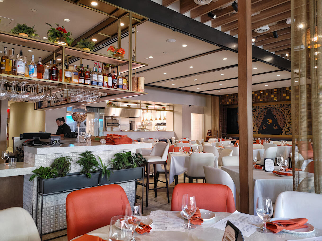 A restaurant with white tablecloths, orange chairs, open kitchen, and shelves displaying bottles at Mahesh's Kitchen.