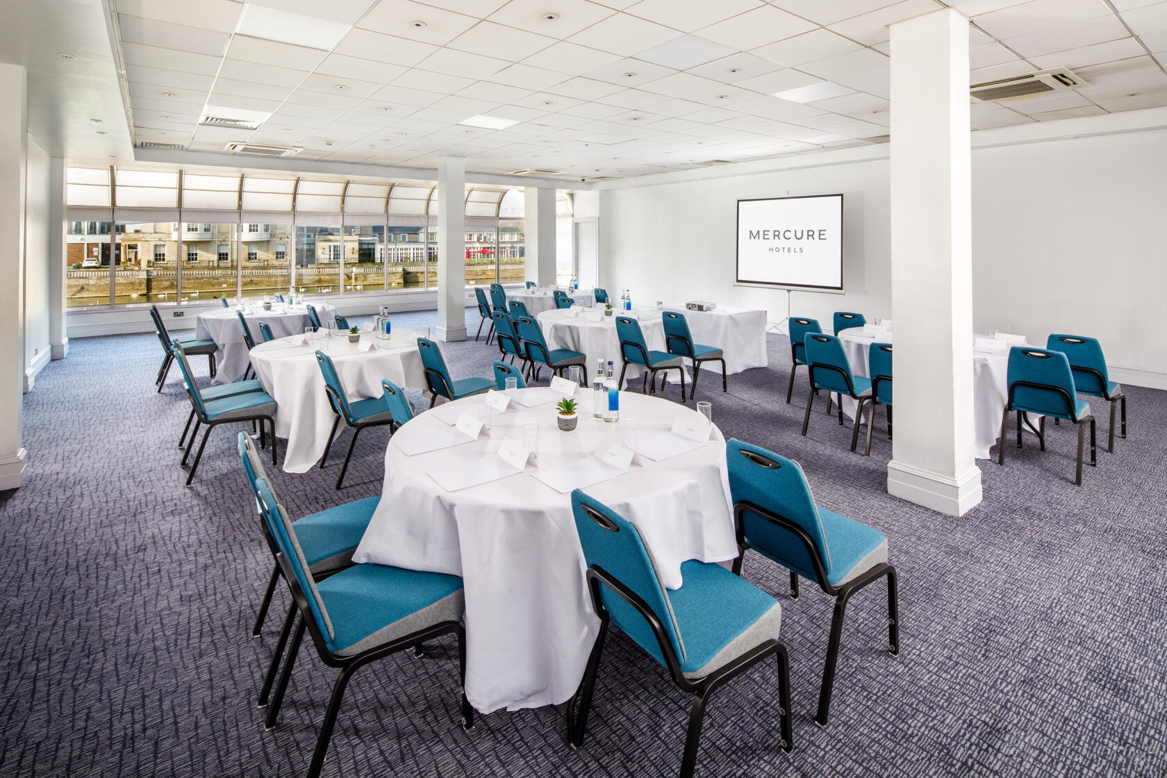 A function room with round tables dressed in white linens, teal chairs, and a projector screen at Mercure Bedford Centre.