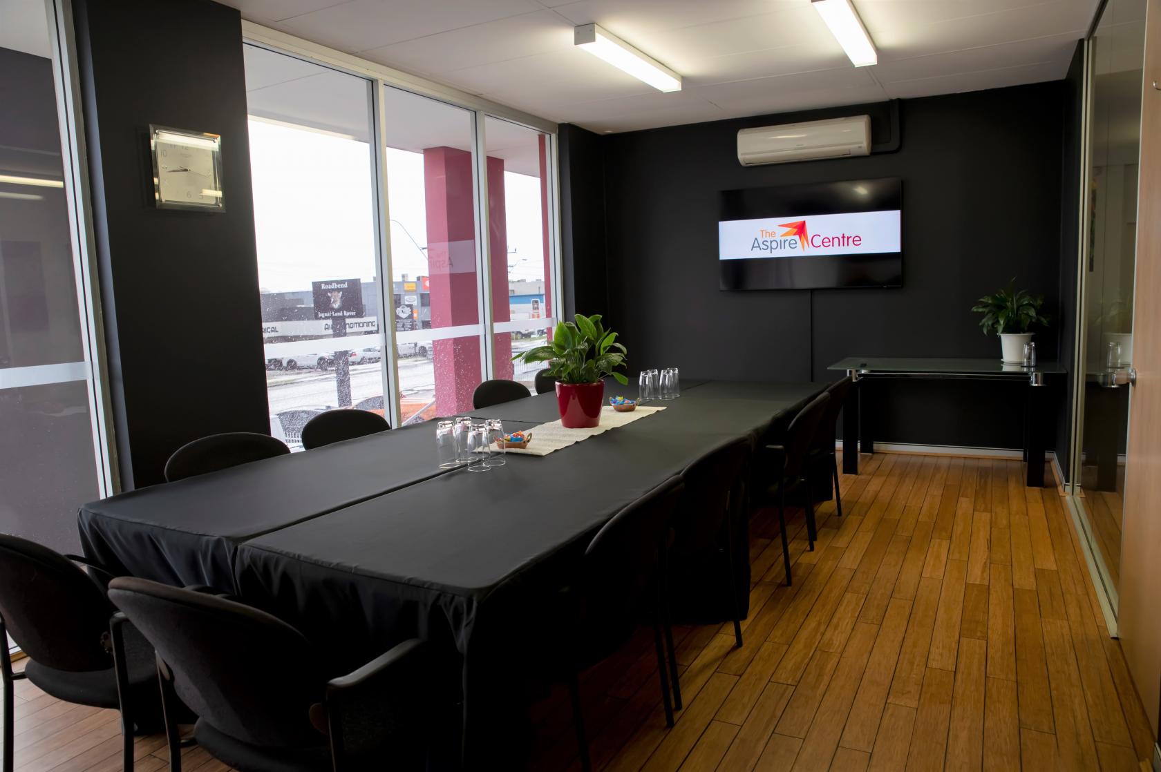 A training room with long tables covered in black linens, a wall-mounted screen, and wood flooring at The Aspire Centre.