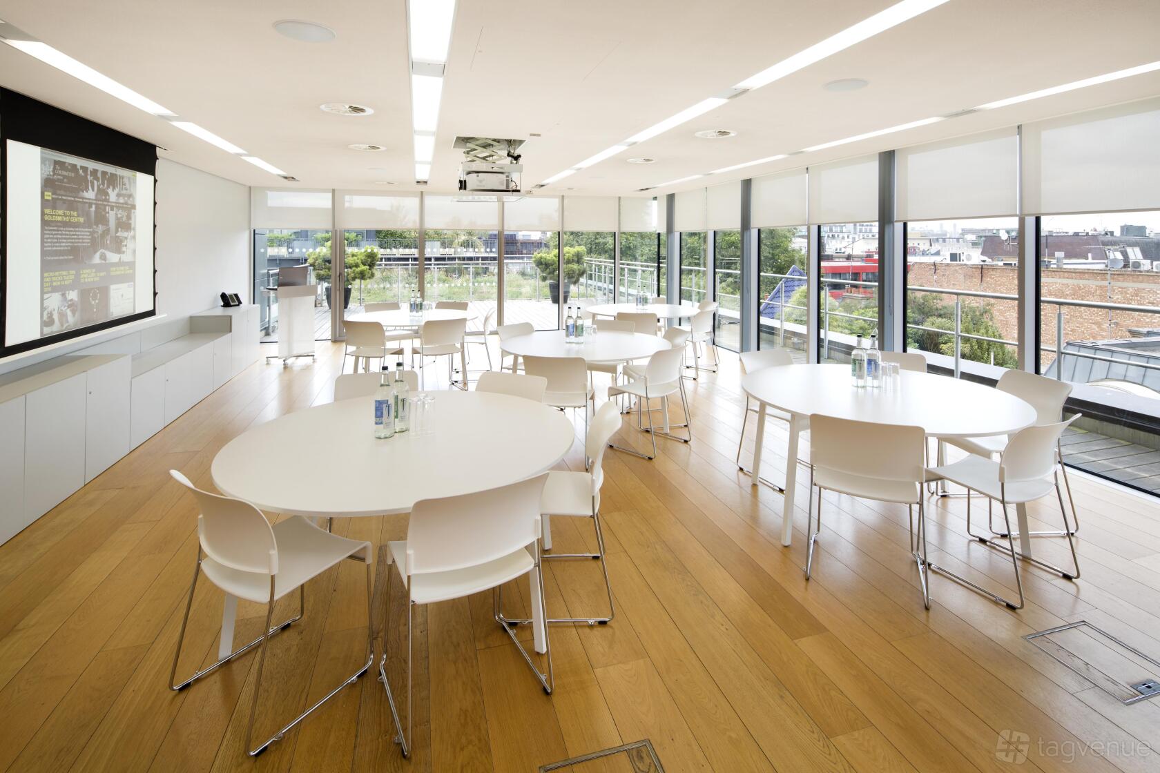 A meeting room with floor-to-ceiling windows, round tables, white chairs, and wood floors at The Goldsmiths' Centre.