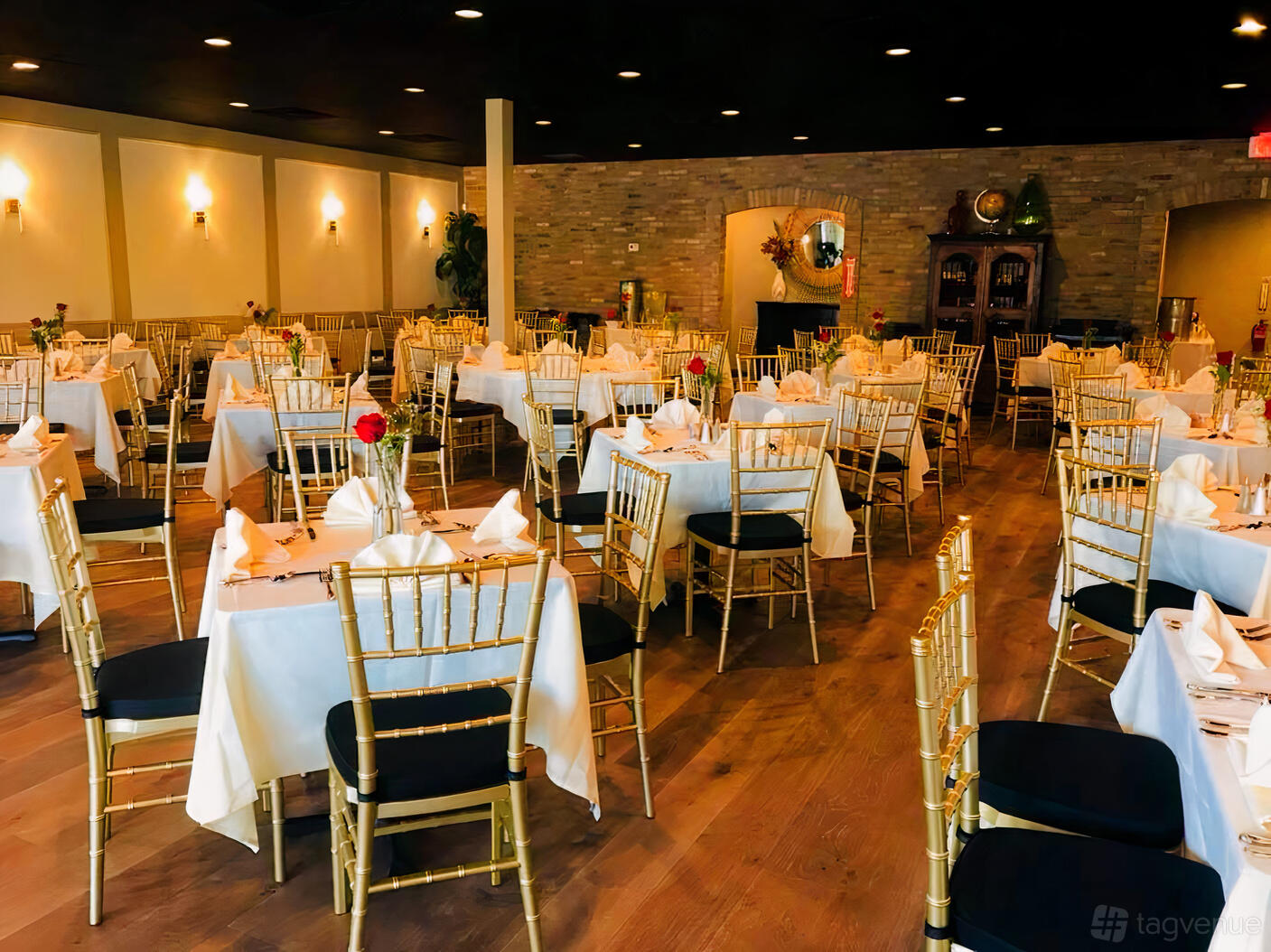 A dining room with round tables draped in white linens, gold chiavari chairs, and brick accent wall at New Season Catering.