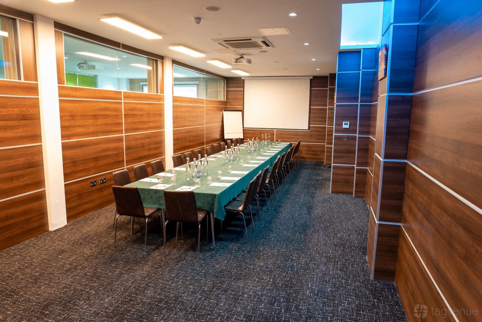 A meeting room with a long rectangular table, green tablecloth, glass walls, and wood paneling at The Wesley Euston Hotel.