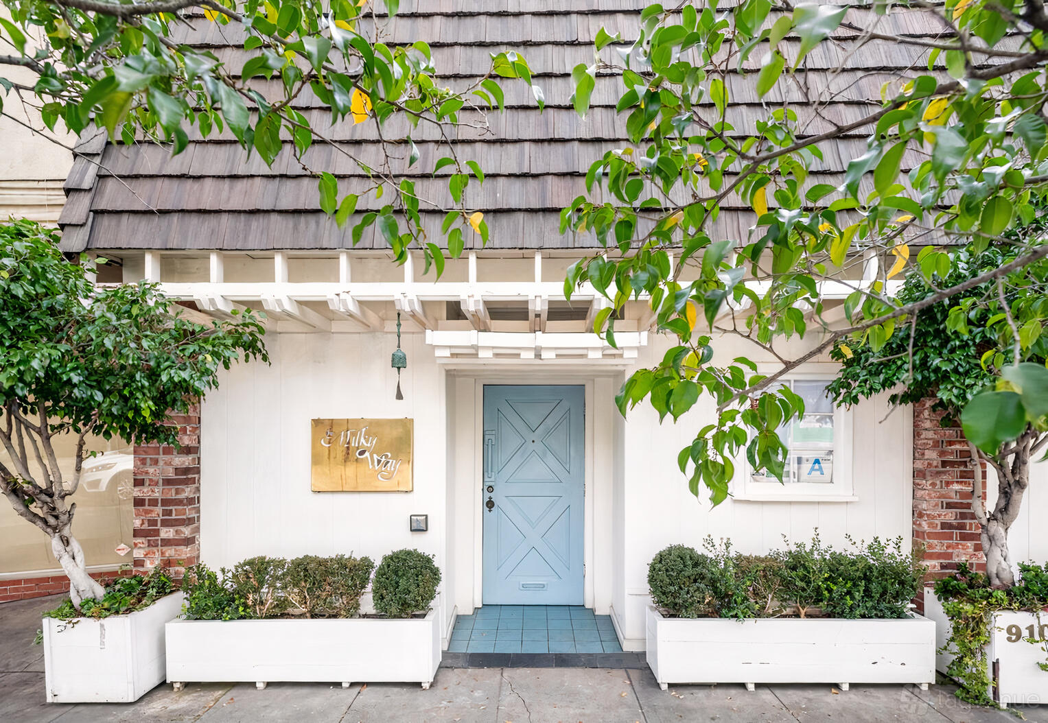 A restaurant entrance with a blue door, white trellis, potted shrubs, and a gold sign at The Milky Way.
