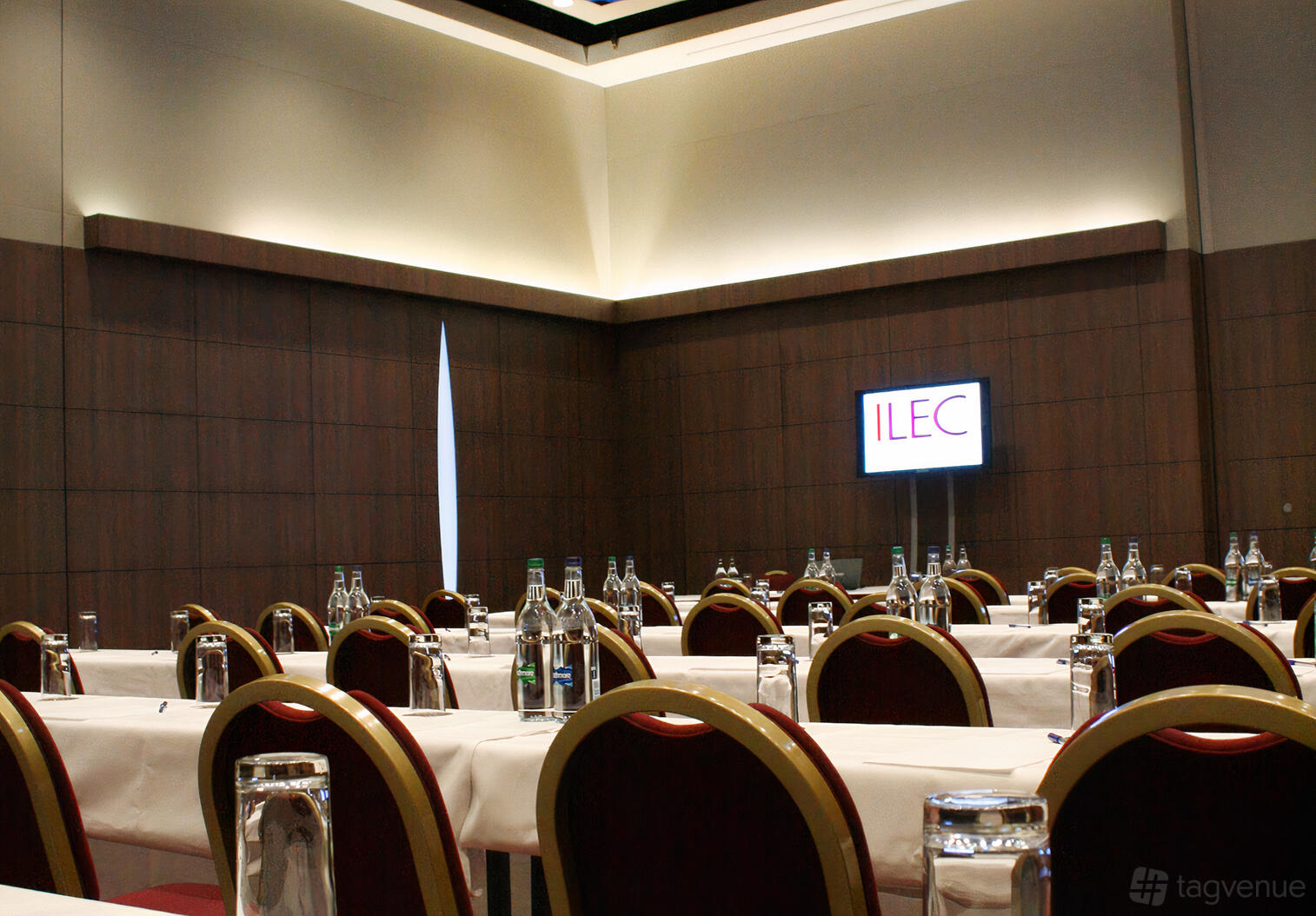 A training room with rows of tables, red cushioned chairs, bottled water, and a wall-mounted screen at Mercure London Earl's Court.