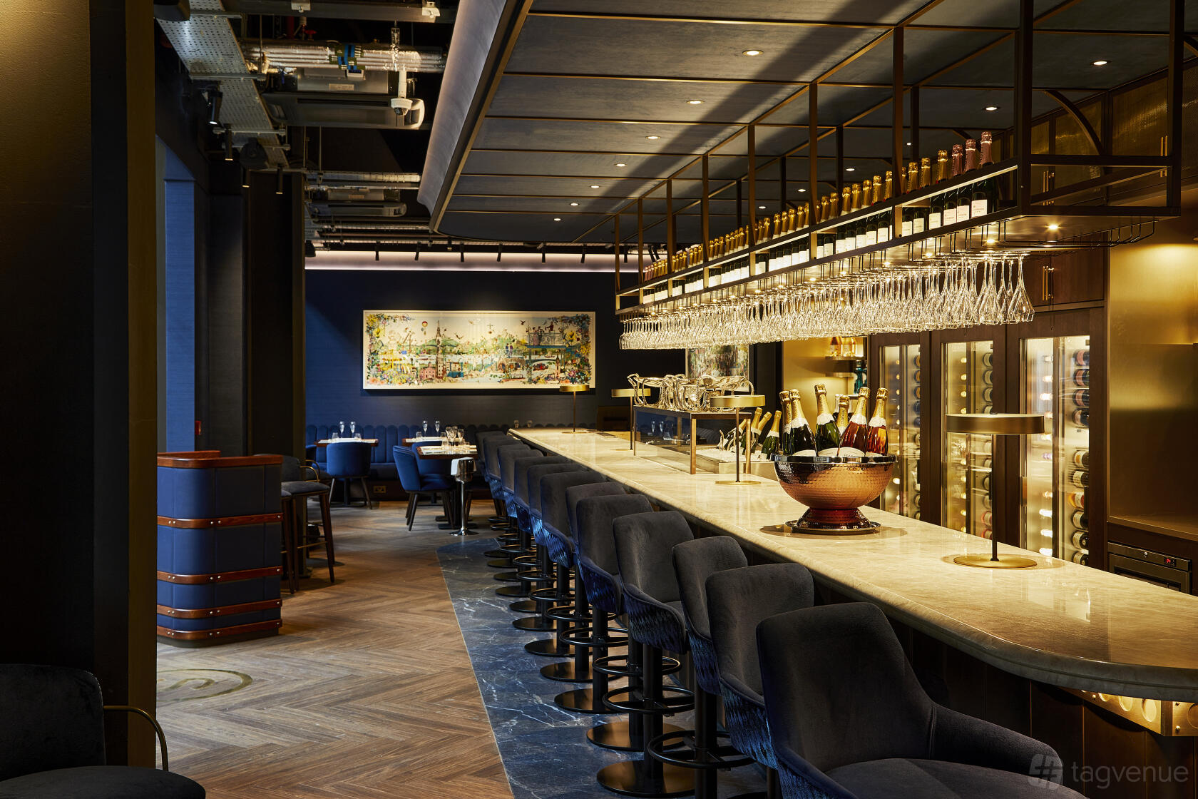 A restaurant with a long marble bar, navy velvet stools, hanging glassware, and wood floors at Searcys Champagne Bar at Battersea.