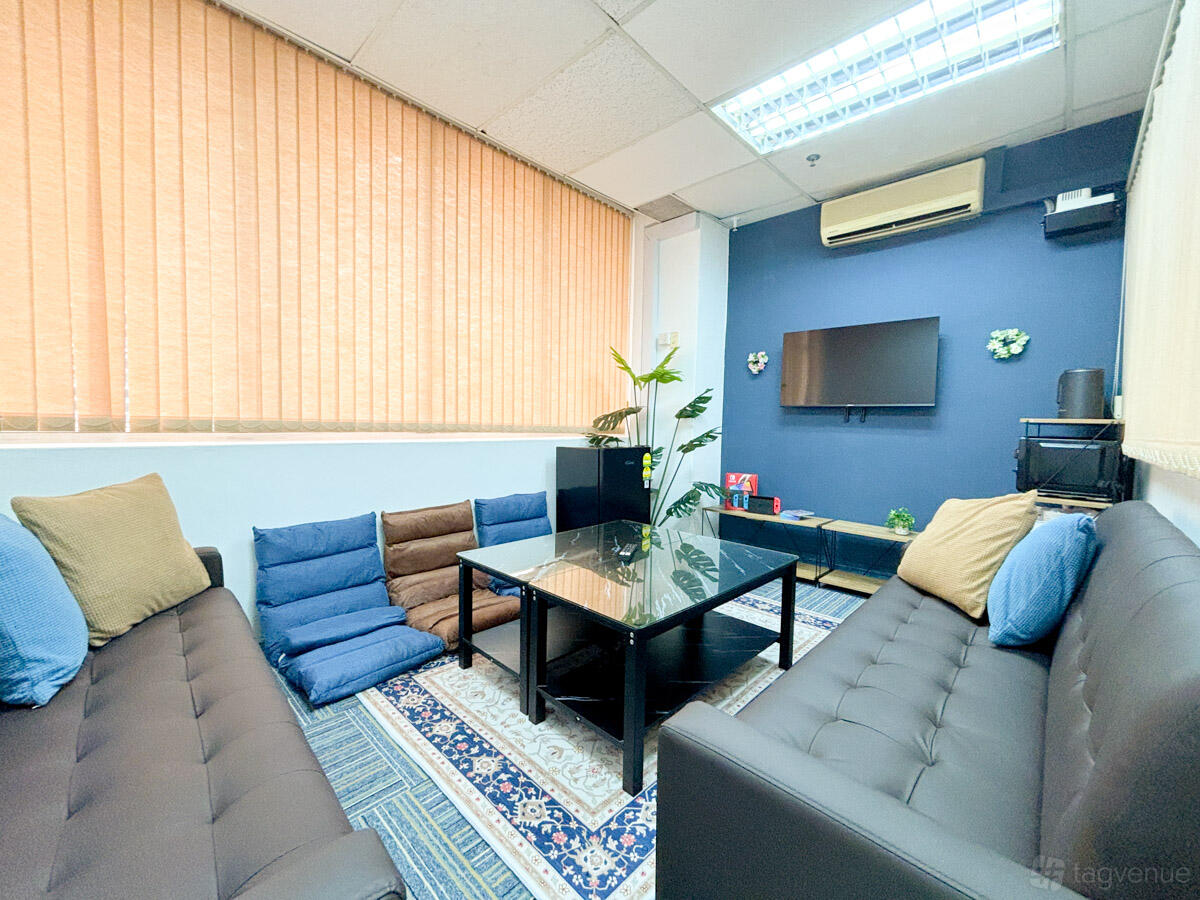 A function room with sofas, a glass coffee table, floor cushions, and a wall-mounted TV at RoomKiosk Tanjong Pagar.