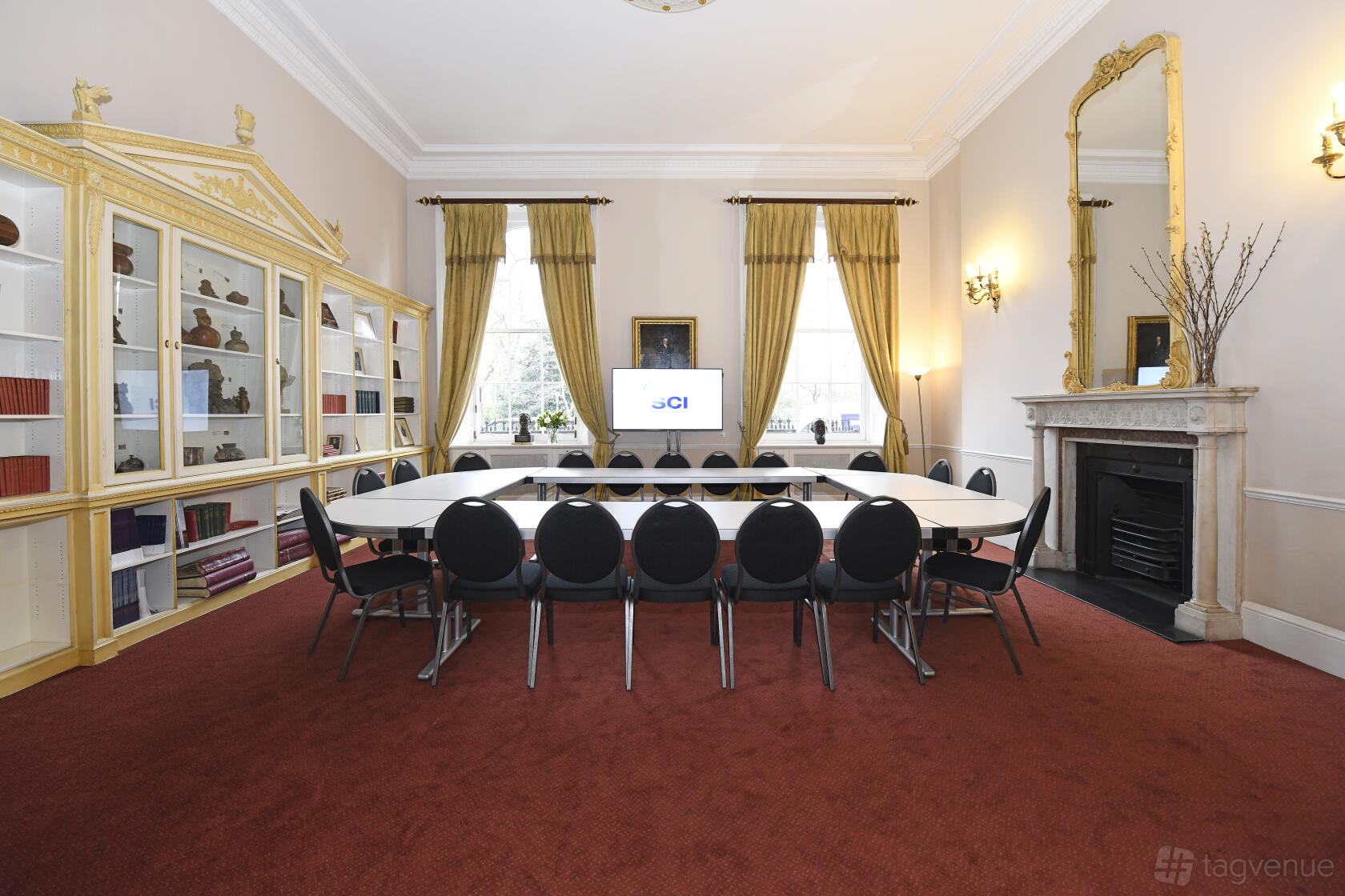 A meeting room with a U-shaped table setup, red carpet, tall windows with gold curtains, and a fireplace at SCI Belgravia.