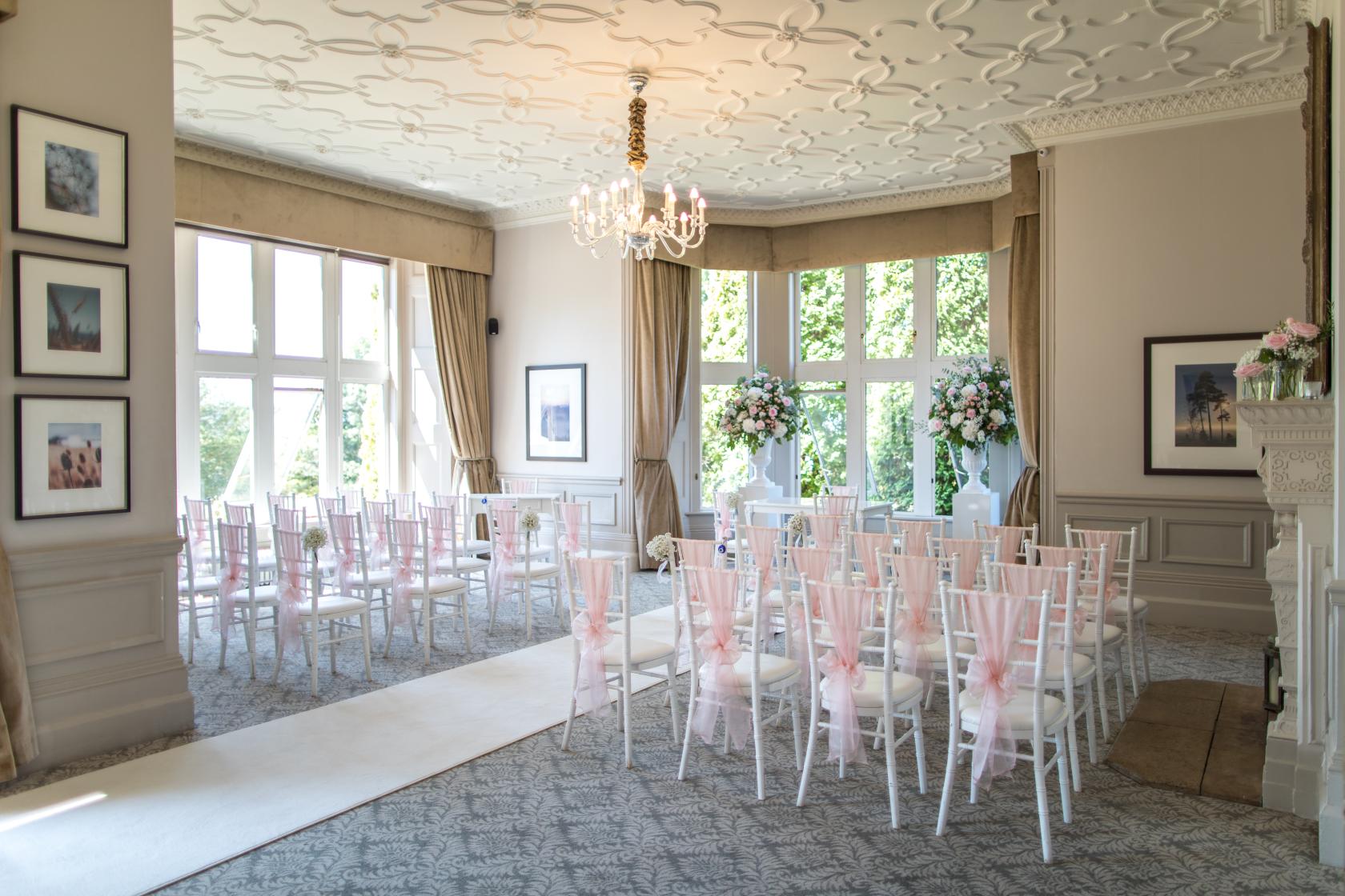 A hotel lounge set up for a ceremony with white chairs, pink sashes, floral arrangements, and large windows at Hartsfield Manor.