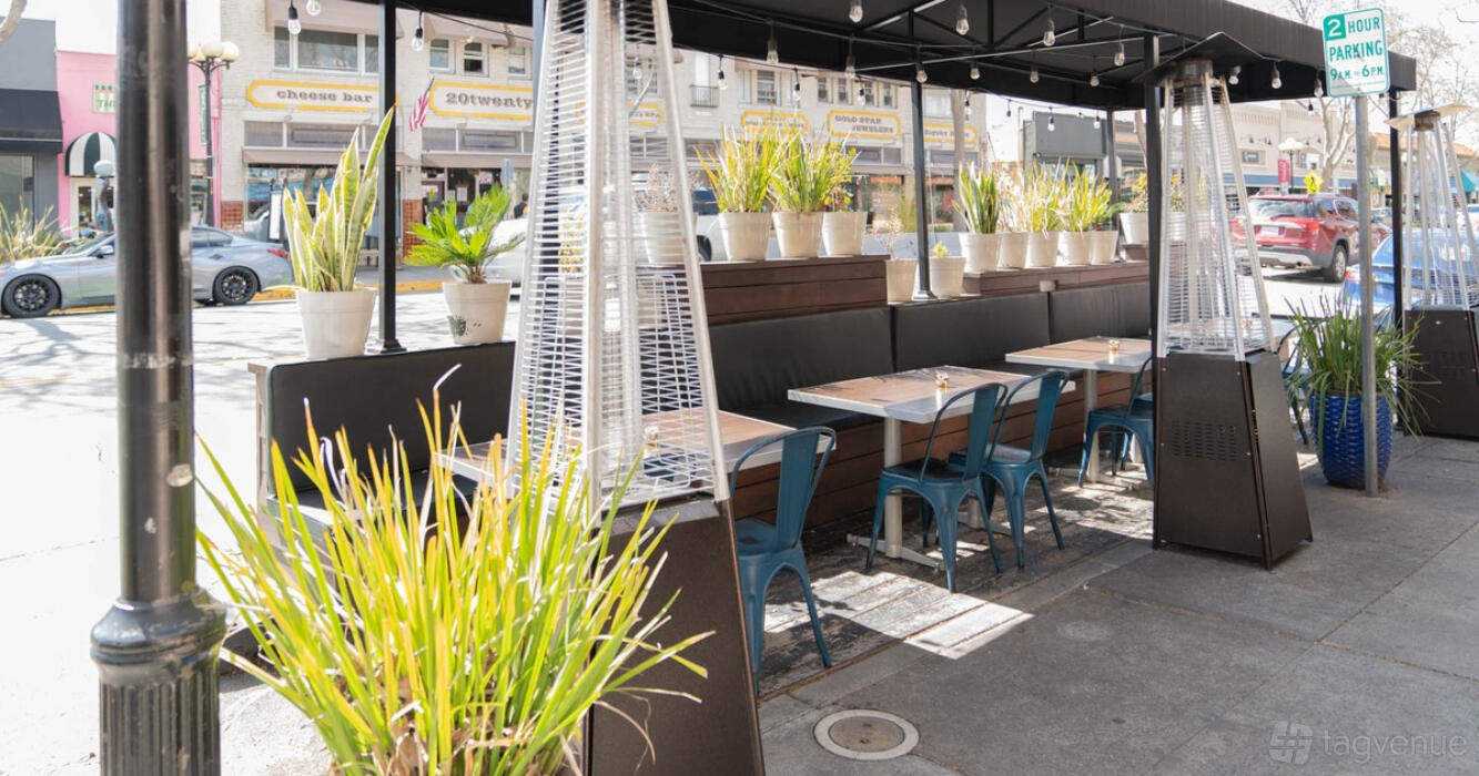 A restaurant patio with potted plants, outdoor heaters, and wooden tables with blue chairs at Arepas Latin Cuisine - San Jose.