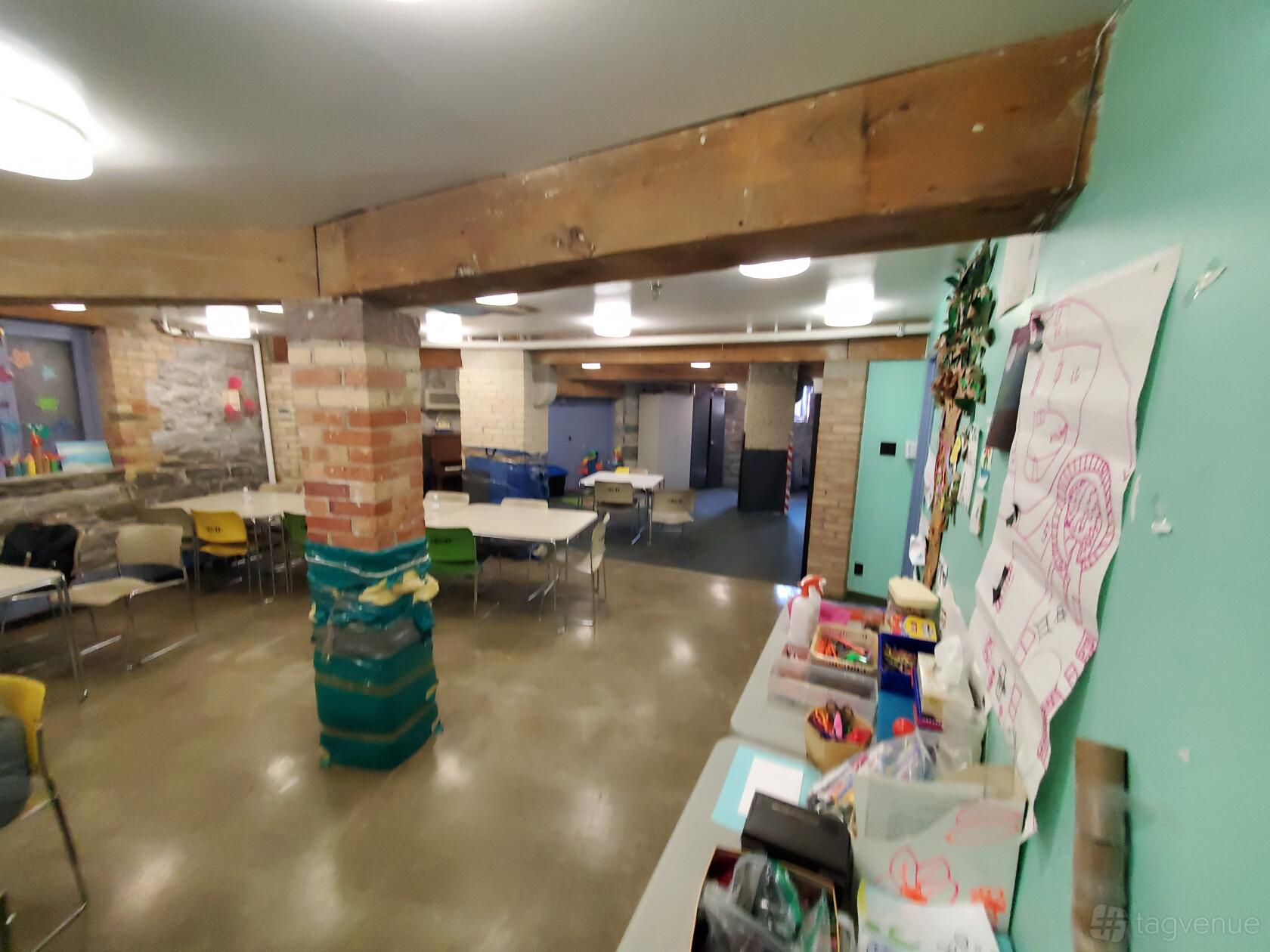 A community centre room with exposed brick columns, children’s artwork, and tables with mixed chairs at Cecil Community Centre.