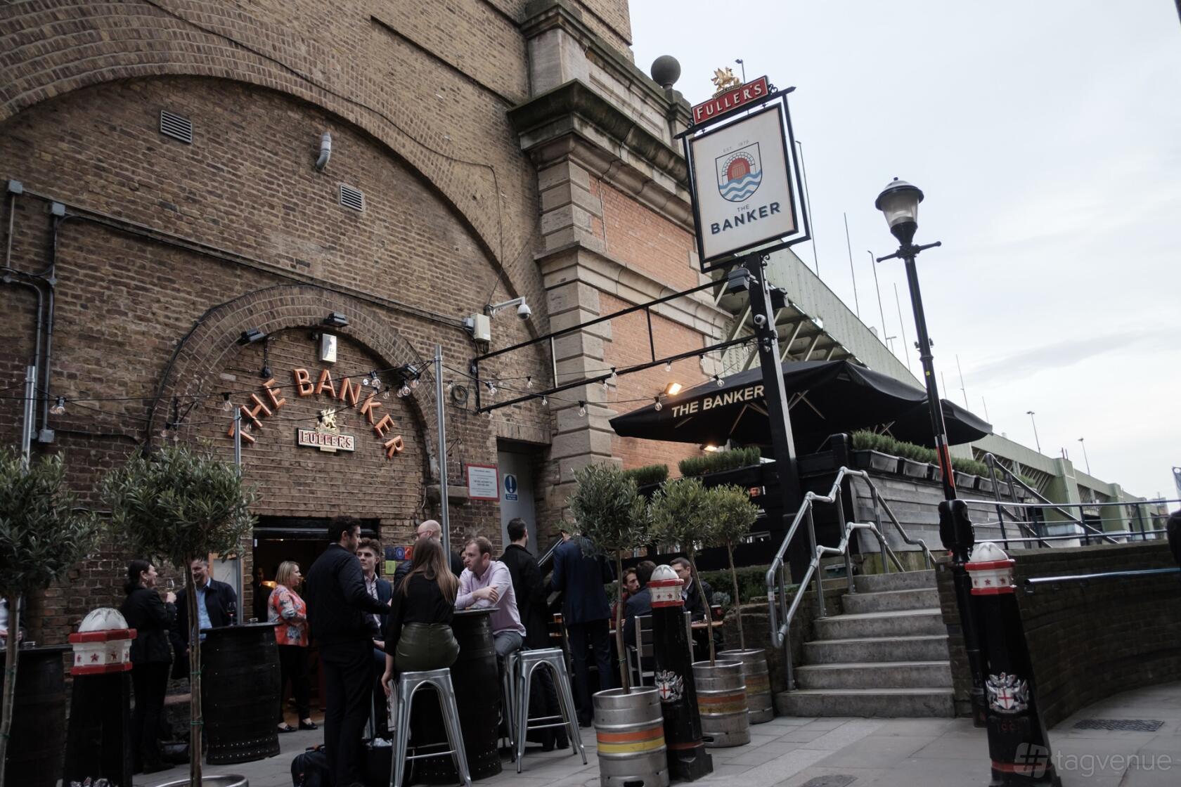 A riverside pub with exposed brick exterior, arched entrance, and outdoor seating beneath The Banker sign