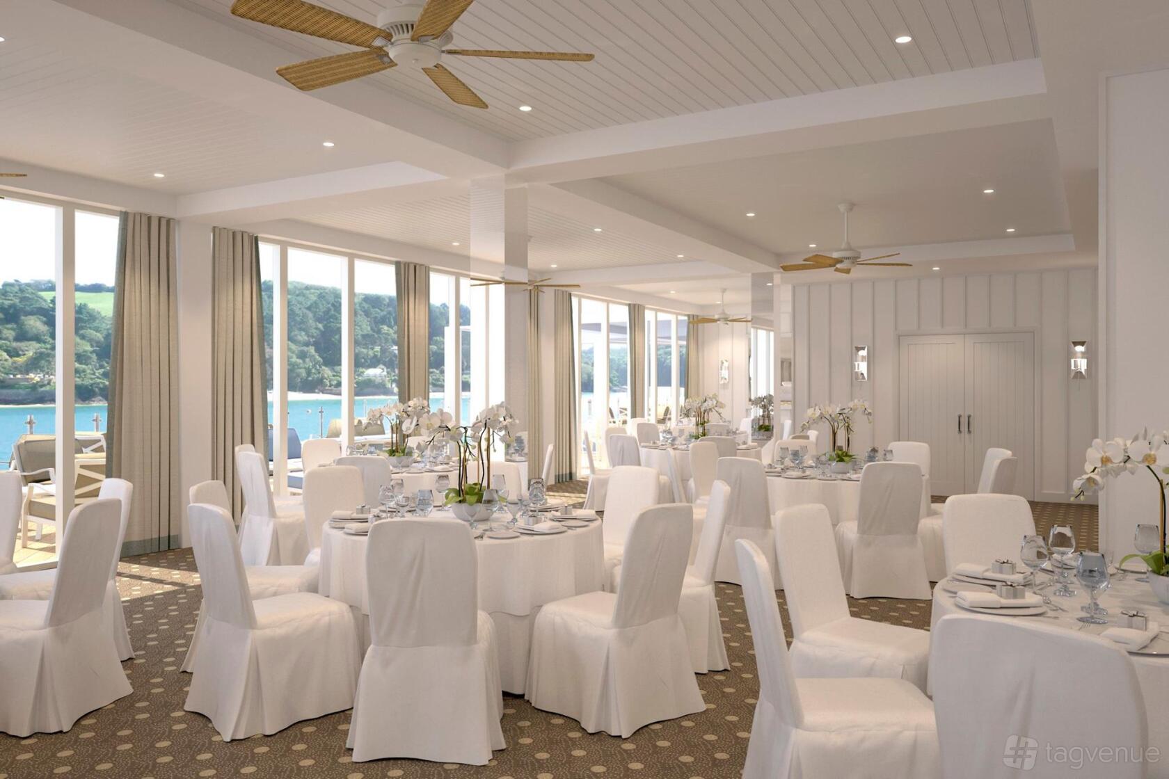 An event space with large windows, round tables draped in white linens, and views of the water at Salcombe Harbour Hotel & Spa.