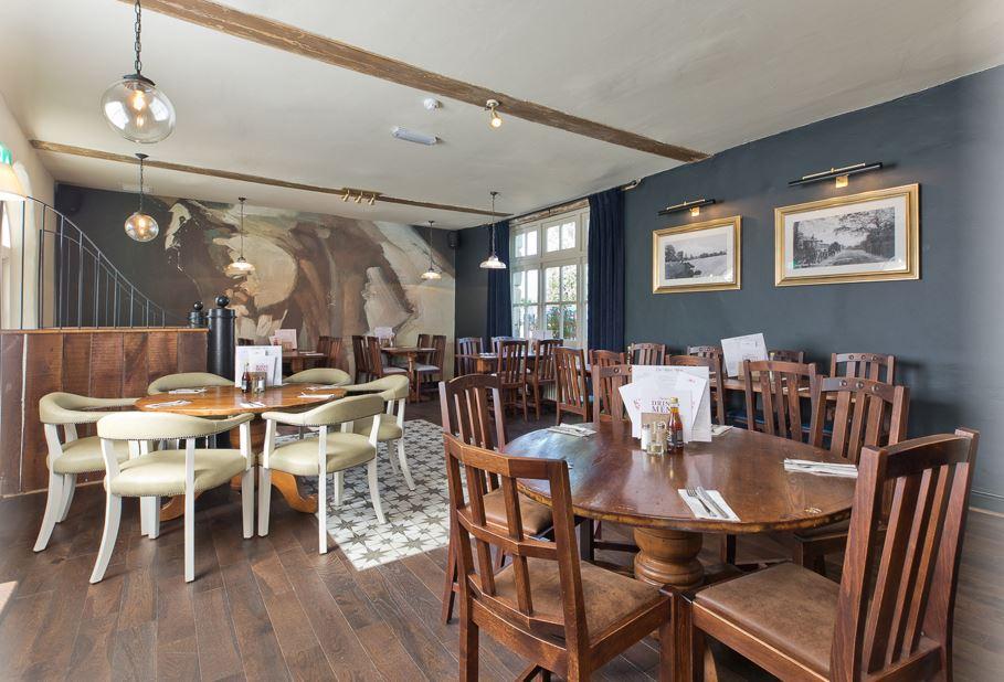 A pub event space with dark wooden tables, cushioned chairs, wall art, and large windows at Danson Stables.