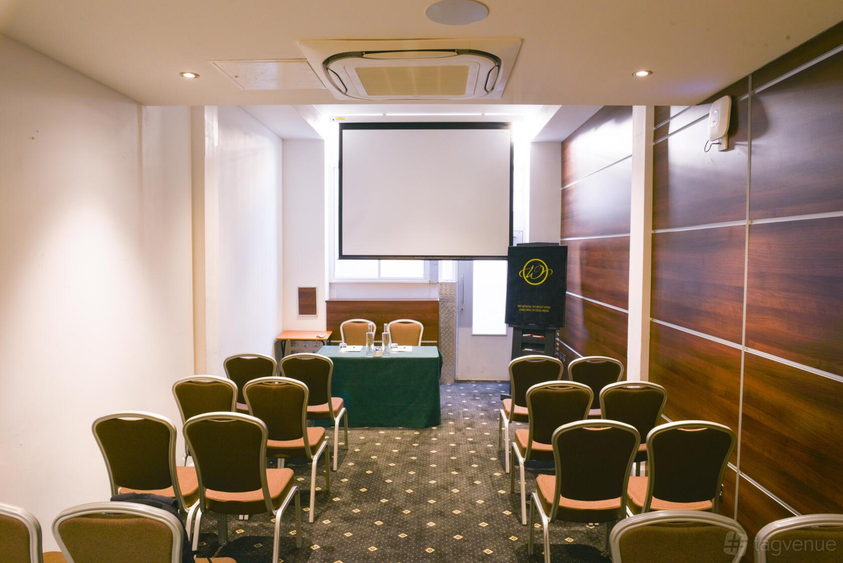 A hotel meeting room with rows of chairs, wood-paneled walls, and a ceiling projector screen at The Wesley Euston Hotel.