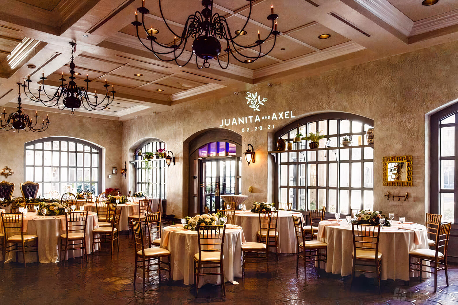 A hall with arched windows, chandeliers, round tables with white tablecloths, and floral centerpieces at The Venetian Terrace.