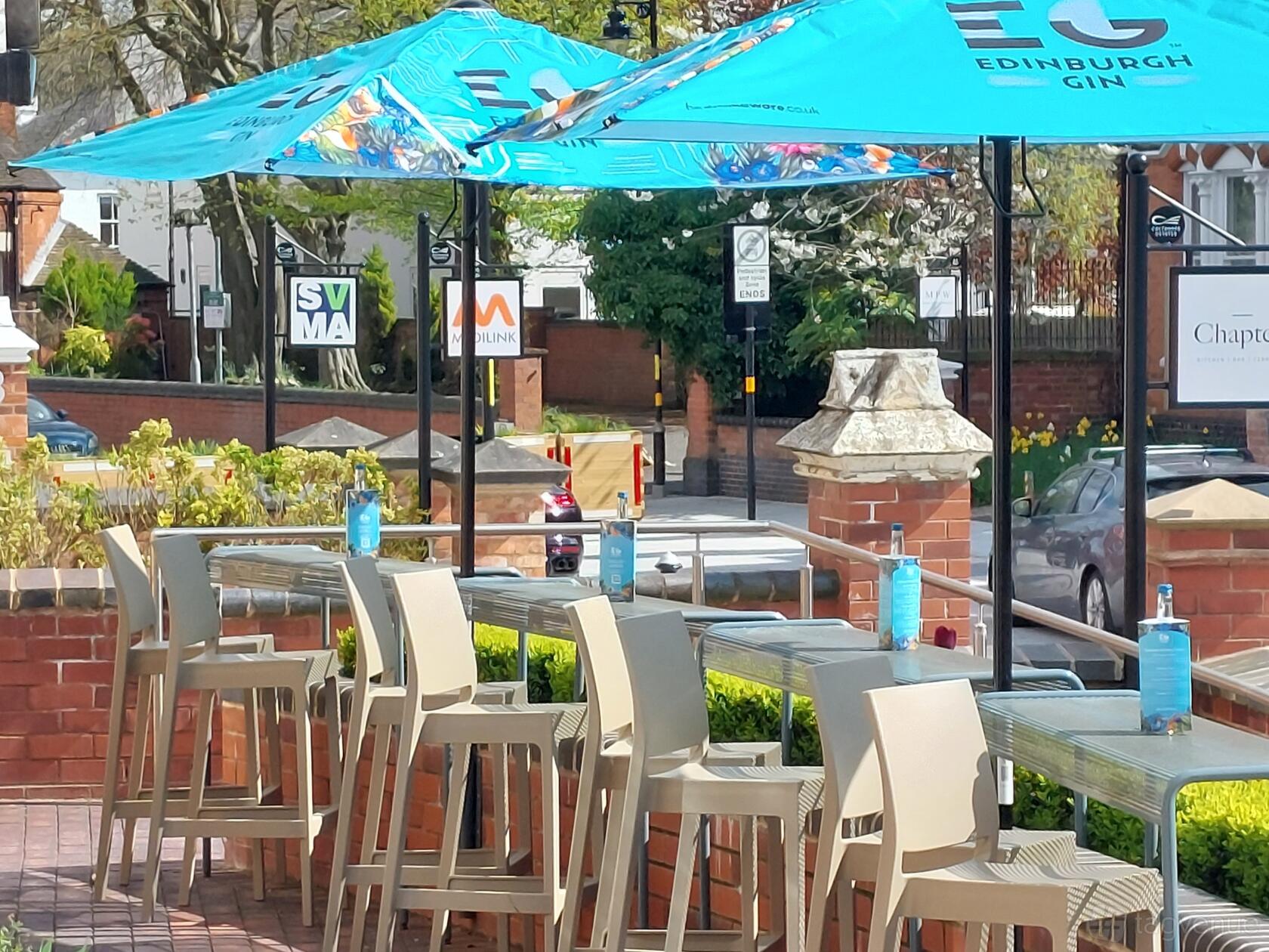 A restaurant terrace with high tables, beige chairs, blue umbrellas, and gin bottles at Chapter Edgbaston.