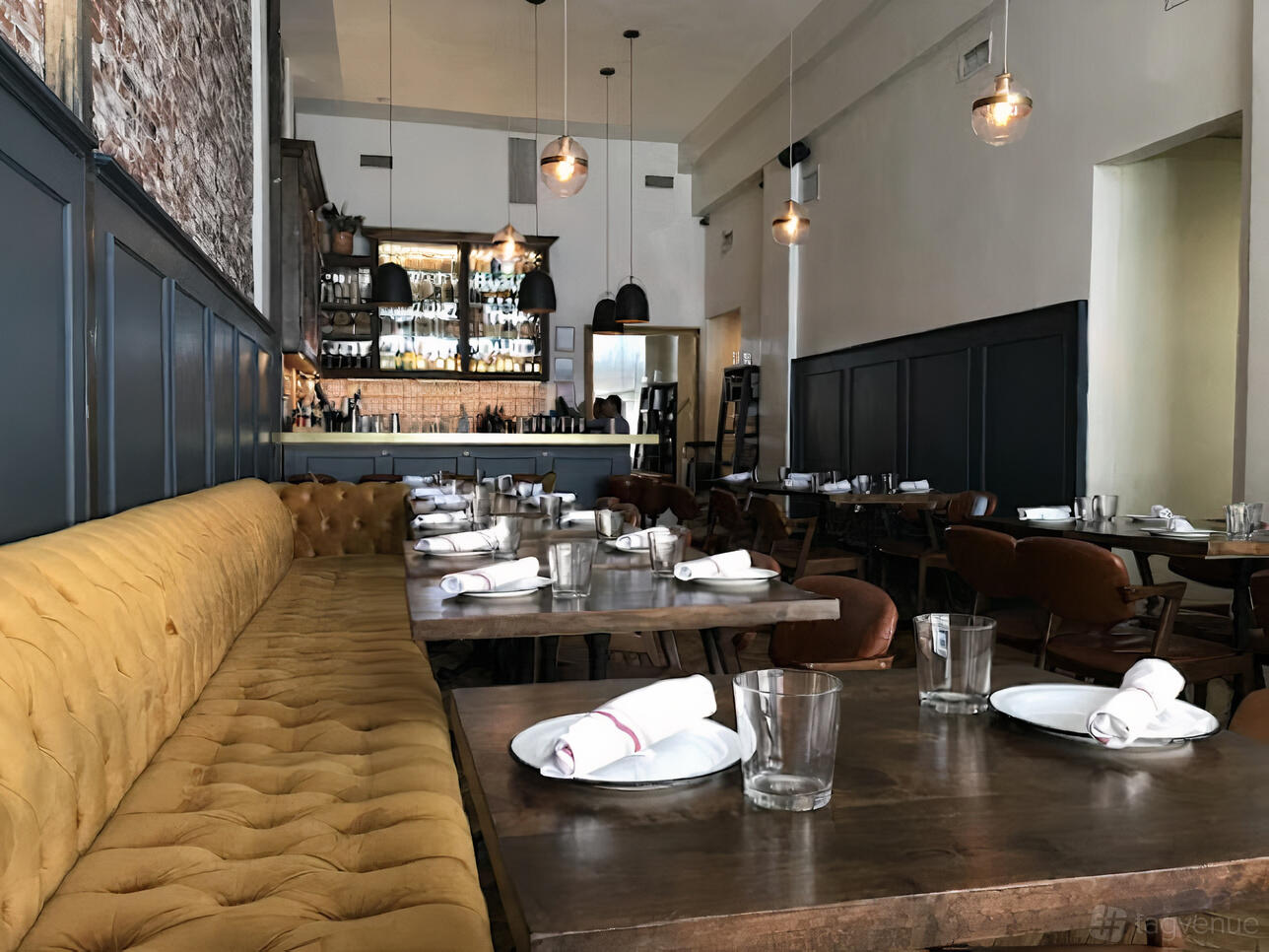 A restaurant interior with a tufted yellow banquette, wooden tables set with plates and napkins, and pendant lighting at Maestro Restaurant.