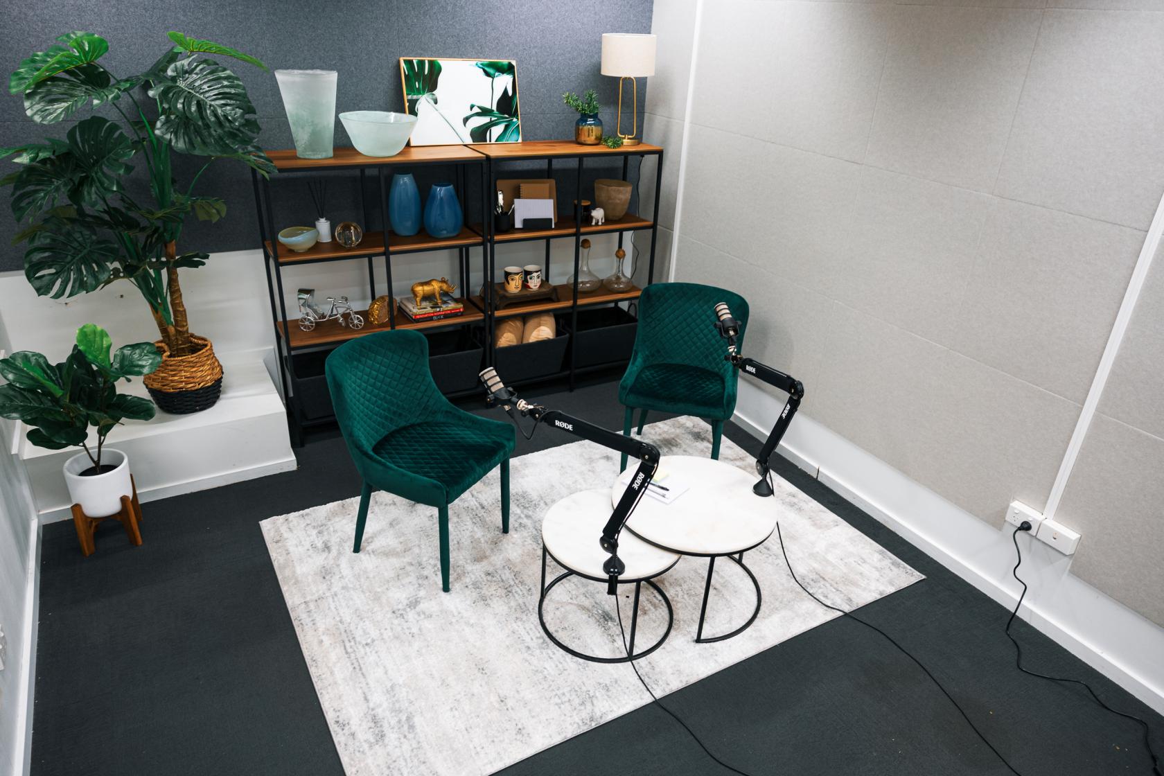A recording studio with two green chairs, a round coffee table, floor plant, and wall shelves at Content Hype Studio.