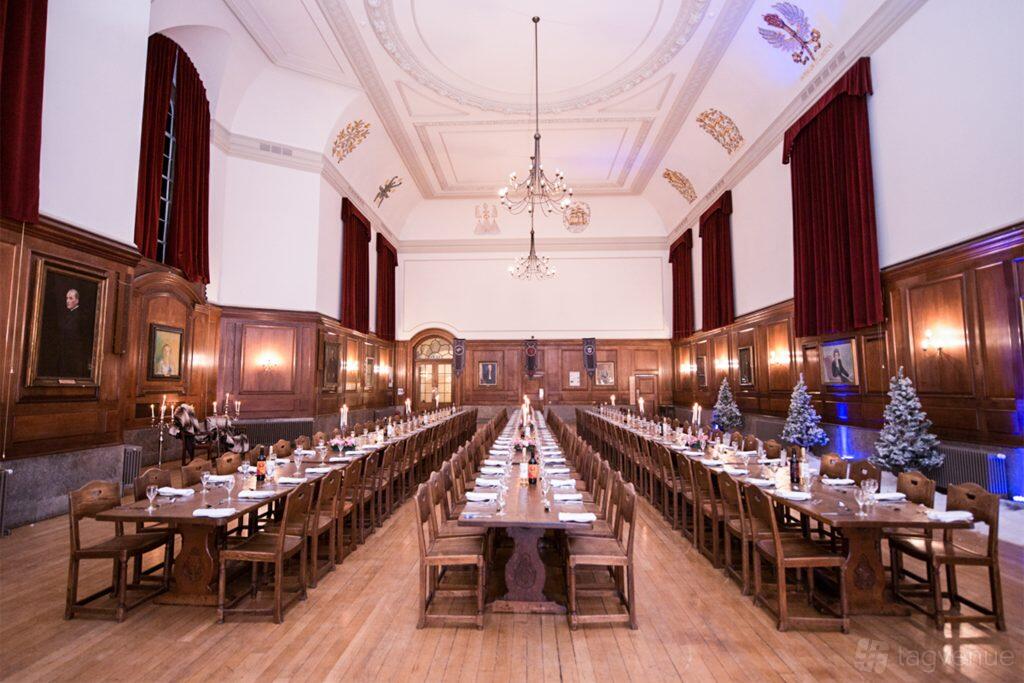 A grand hall with wood-paneled walls, chandeliers, long banquet tables, and red curtains at Goodenough House.