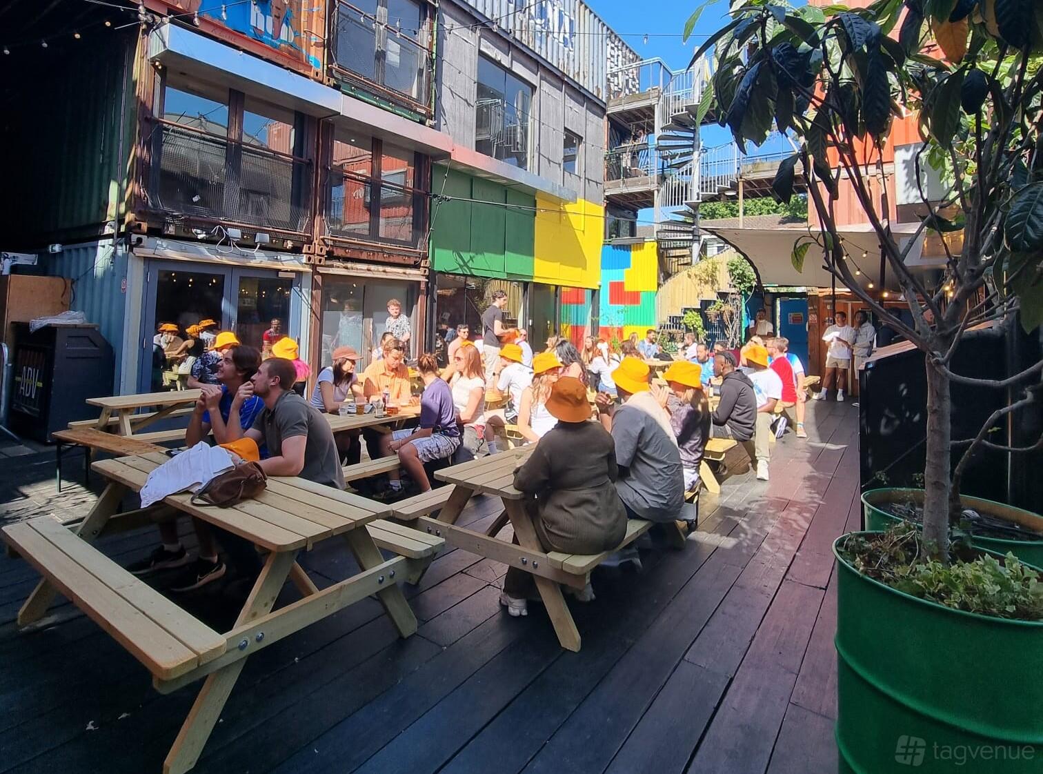 An outdoor entertainment space with wooden picnic tables, colorful shipping containers, and potted trees at Pop Brixton.