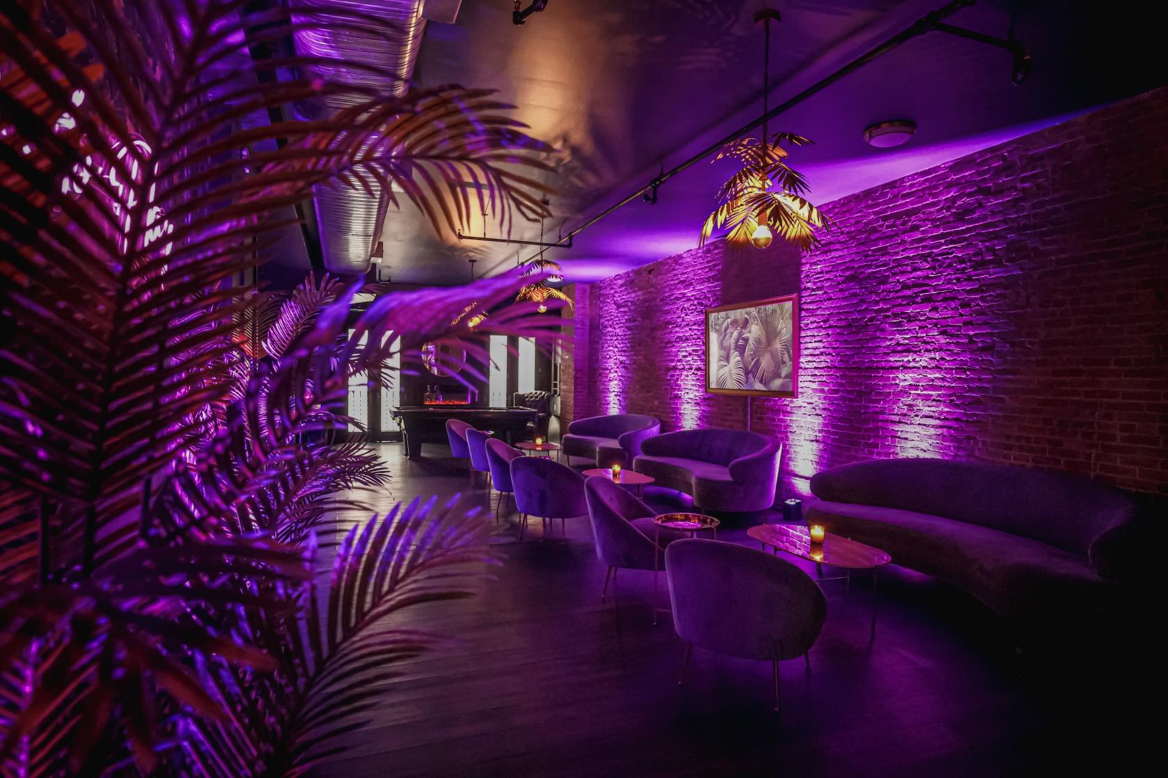 An event loft with exposed brick walls, purple ambient lighting, velvet sofas, and small round tables at Upscale Full Service Loft in Flatiron.
