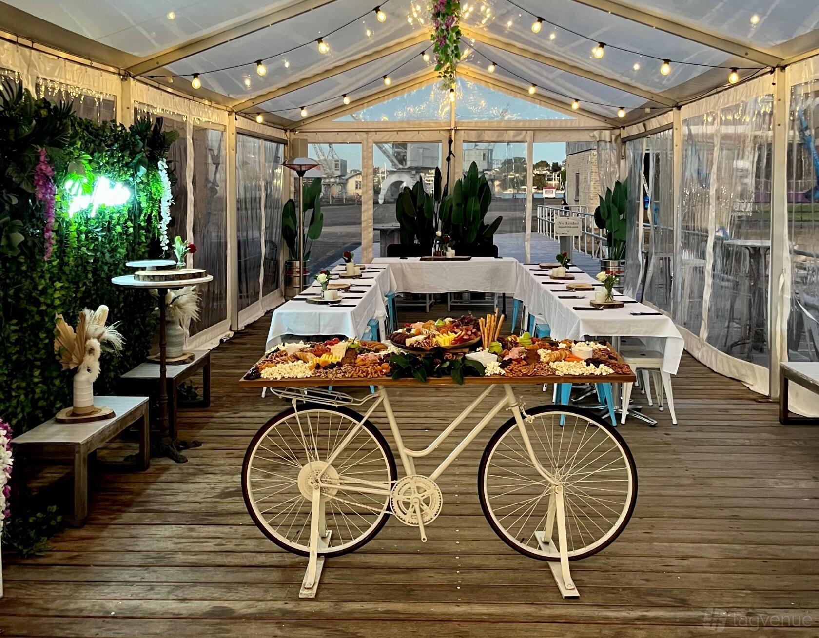 A marquee cafe with long banquet tables, clear tent walls, string lights, and a grazing table on a bicycle cart at Marina Cafe & Bar.