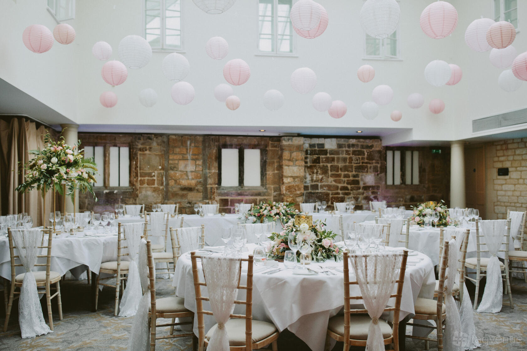 An event space with round tables draped in white linens, wooden chairs, and paper lanterns hanging from a skylight at Ellenborough Park.