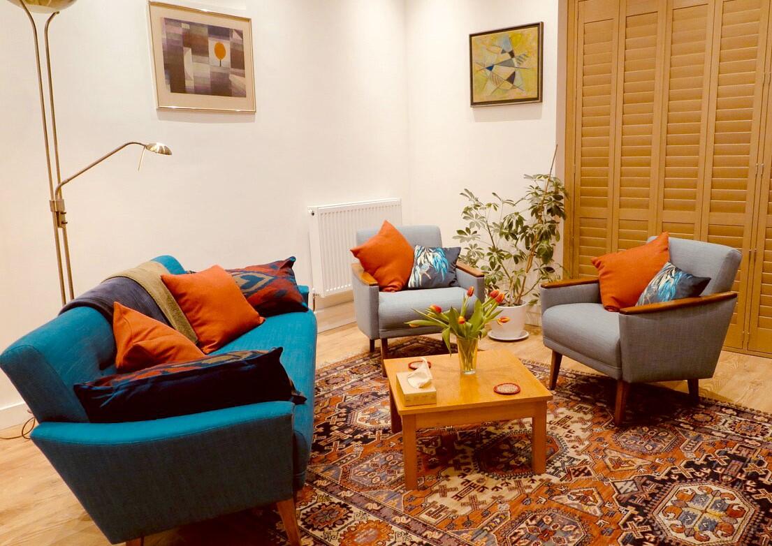 A therapy room with blue armchairs, a matching sofa, patterned rug, and plants at Delancey Therapy Rooms.