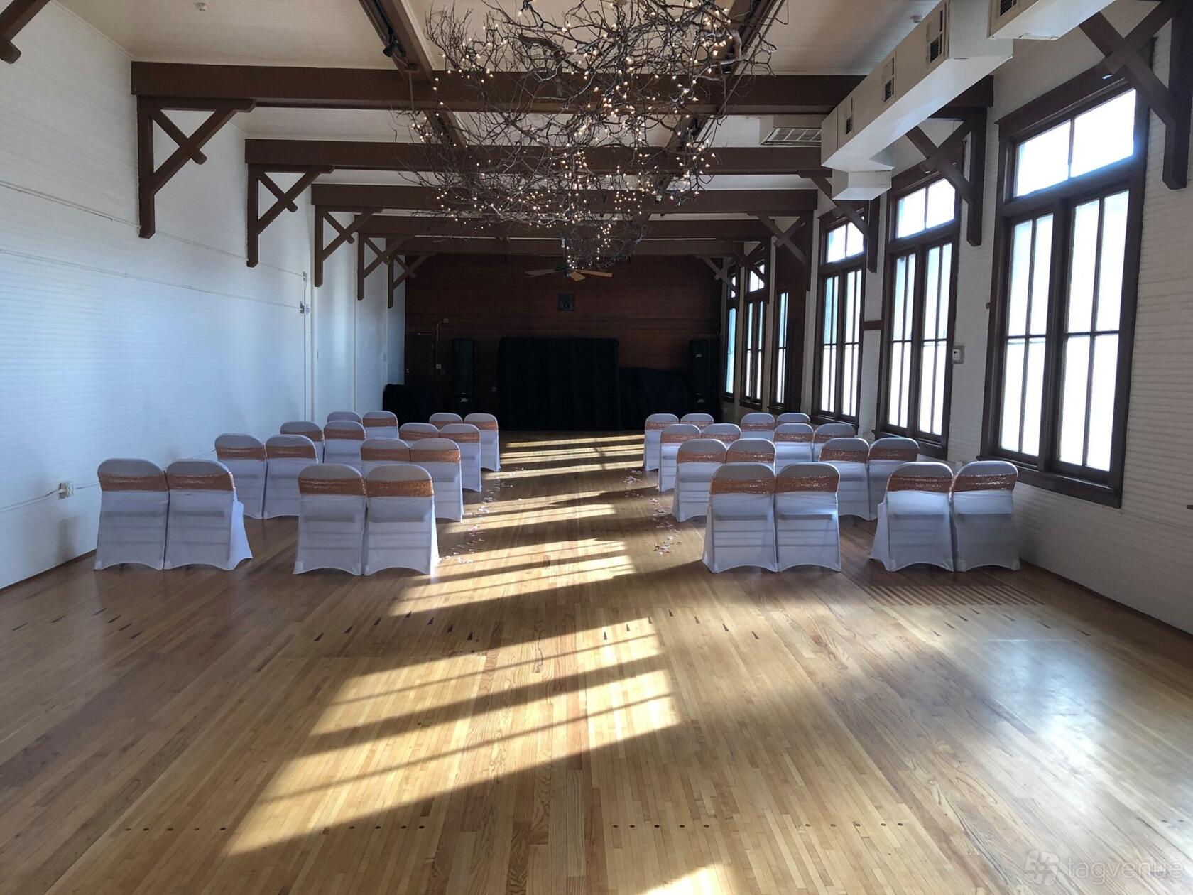A bowling alley event space with wooden floors, rows of white-covered chairs, and large windows at Sons of Hermann Hall.