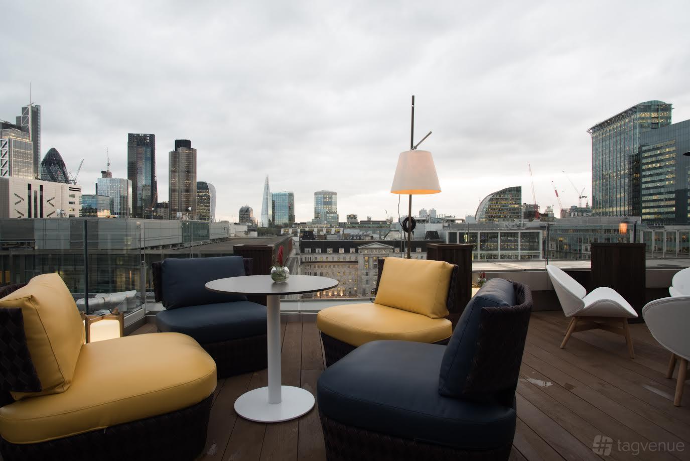 A rooftop terrace bar with city views, cushioned lounge chairs, and small round tables at Aviary Rooftop Bar & Restaurant.