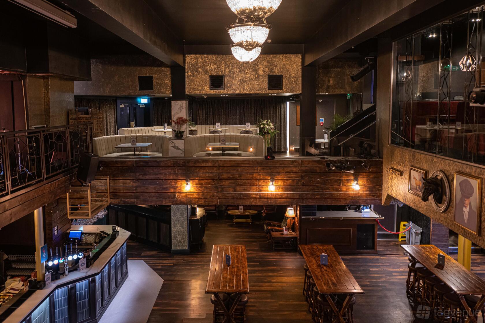 A bar with wooden tables, exposed brick accents, chandeliers, and a mezzanine at Peaky Blinders Manchester.
