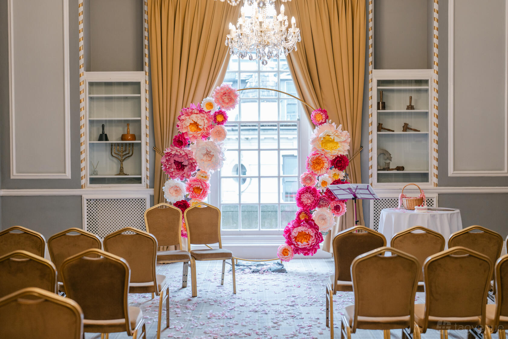 An event space with gold chairs, a large arched window, chandelier, and floral ceremony arch at Manchester Hall’s The Library.