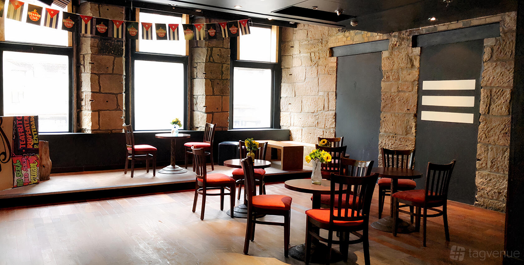 A function room with exposed stone walls, wooden floors, small tables with red-cushioned chairs, and large windows at Mango Latino.